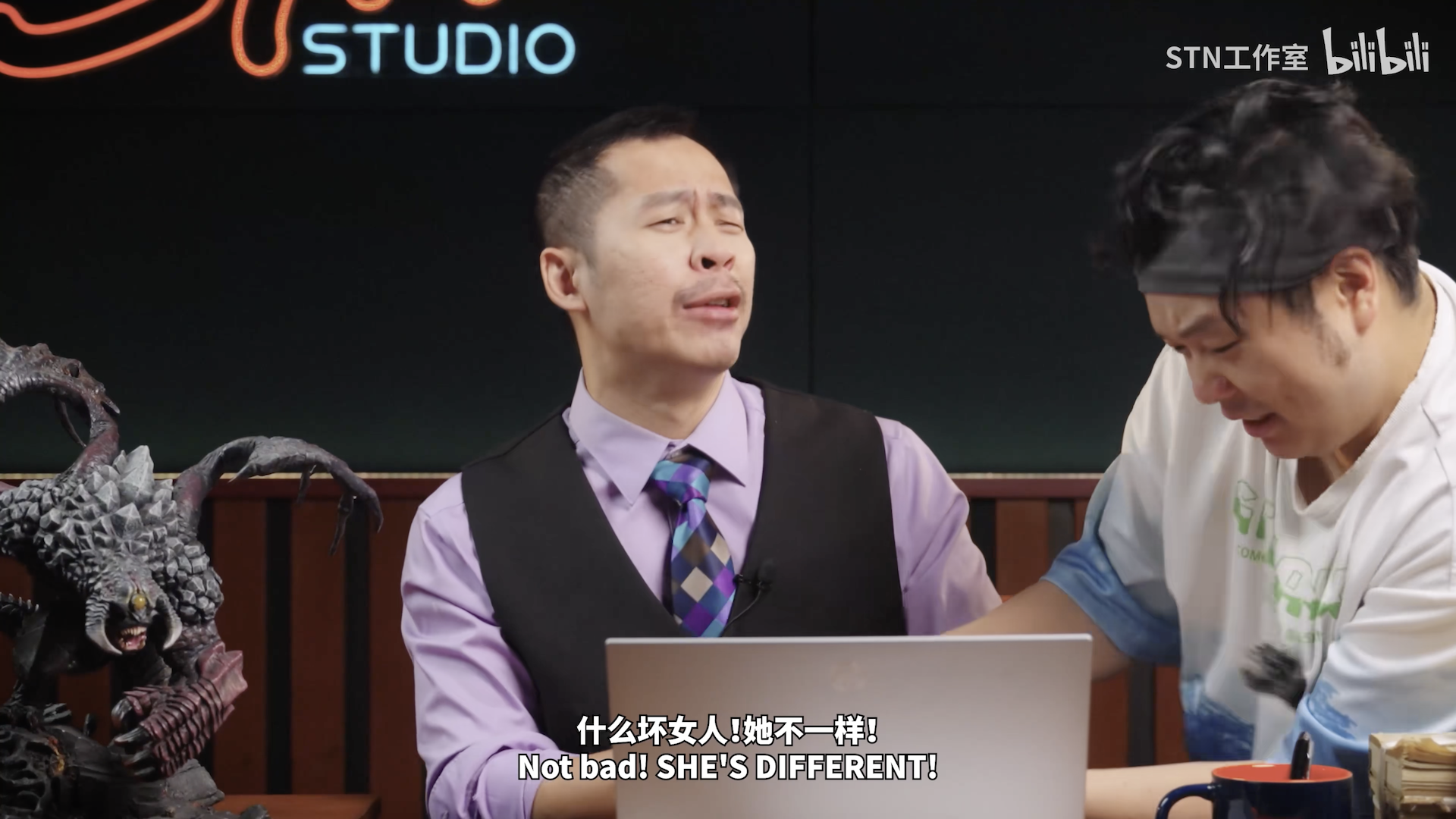 一位穿着紫色衬衫和黑色马甲的男子表情夸张,似乎在激动地说话,旁边另一位穿白色 T 恤的男子试图阻止黑色马甲男子。两人坐在桌前,前方有一台笔记本电脑,左侧有一只精致的肉山手办。底部有字幕:「什么坏女人!她不一样!」