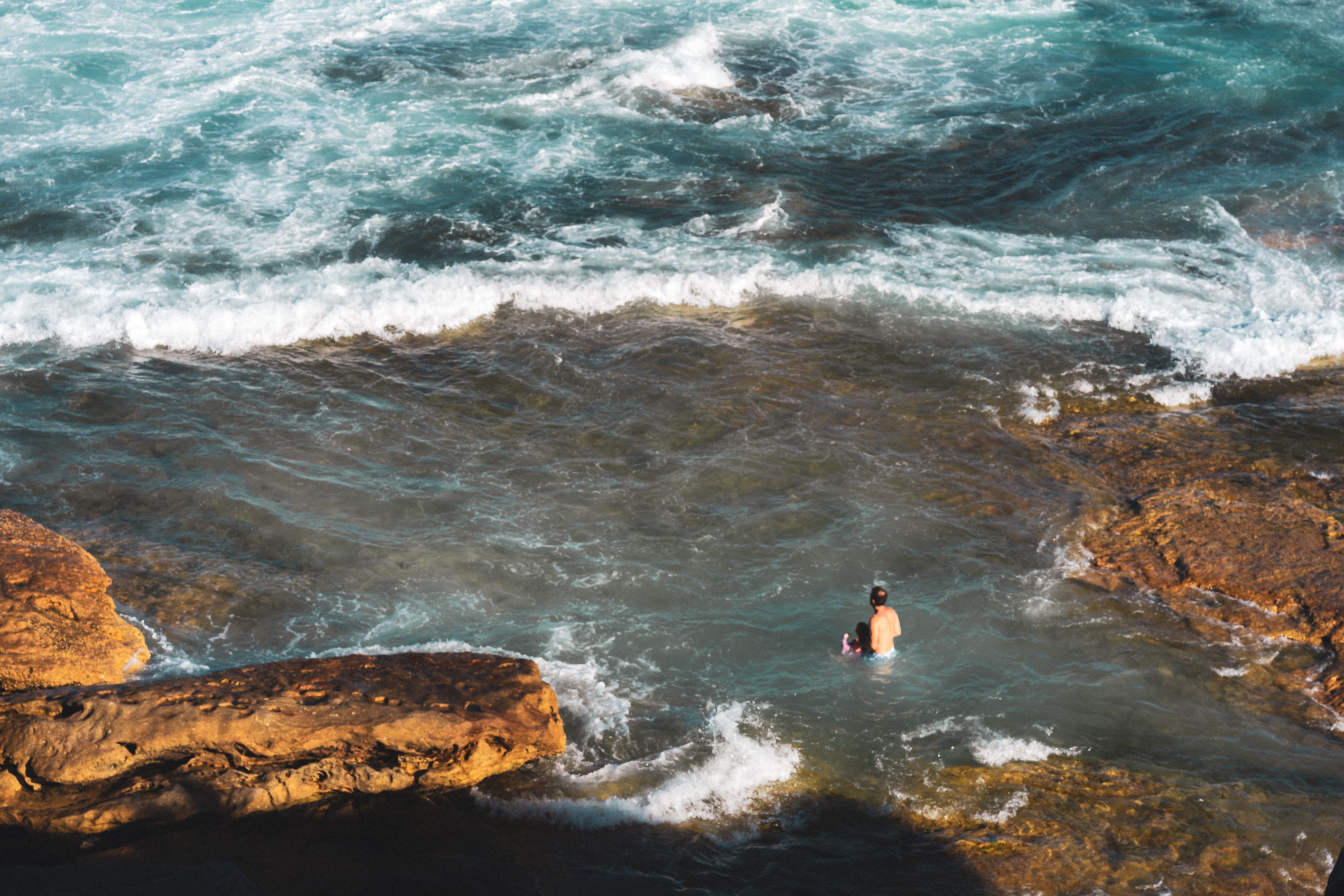 The photograph depicts a vibrant coastal scene with a man and a small child wading in a shallow, rocky pool at the edge of the sea. The water is clear, showcasing the rough textures of submerged rocks, while gentle waves ripple around them. Surrounding the pool are warm, sunlit rock formations with their surfaces worn and smooth, contrasting with the churning turquoise ocean in the background. The movement of the water adds dynamic energy, while the presence of the man and child provides a sense of scale and human connection to the rugged beauty of the natural environment.