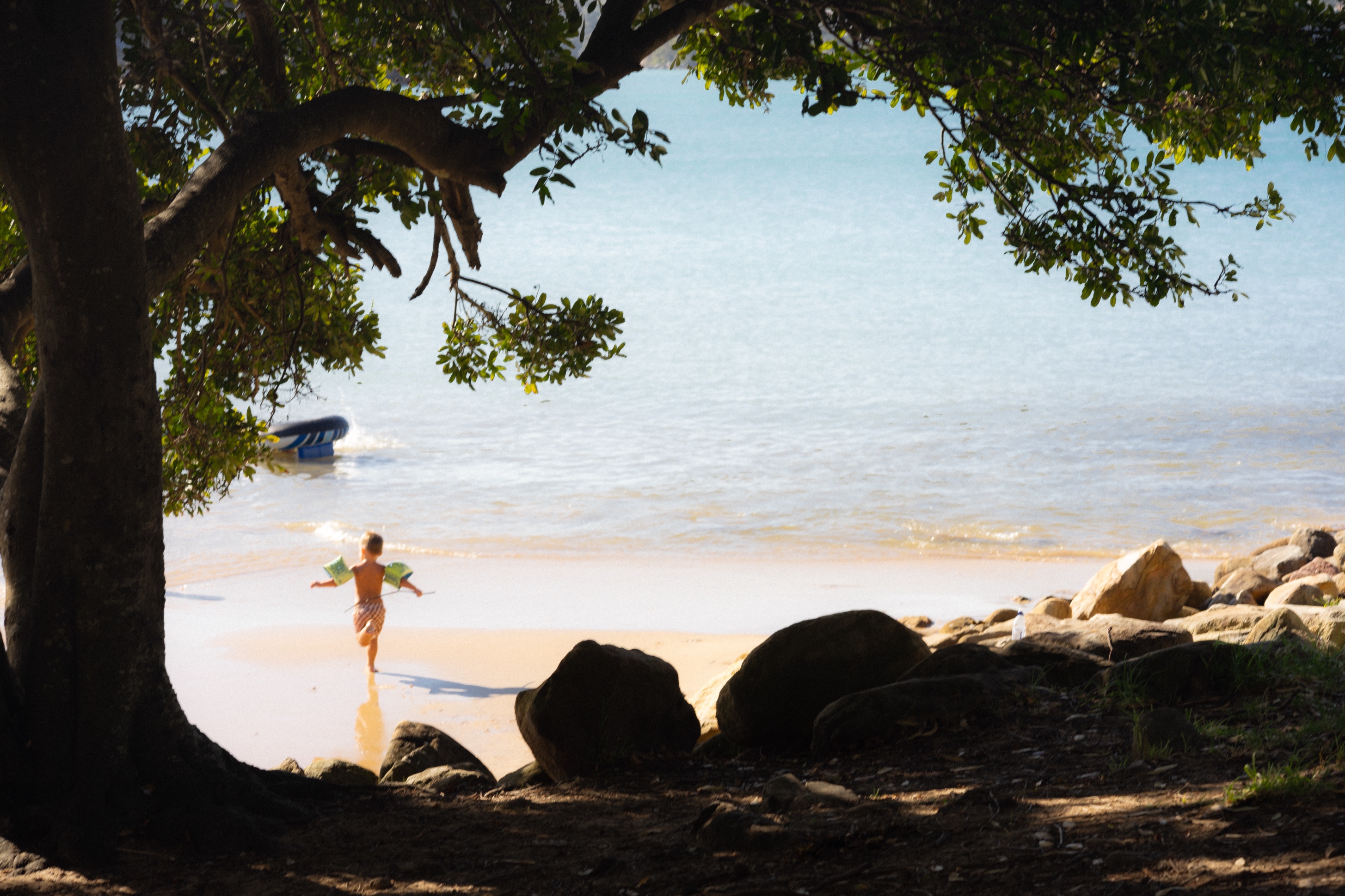 The photograph captures a serene beach scene, framed by the dense, dark foliage of trees in the foreground. A young child, wearing inflatable armbands and light swimwear, is seen running enthusiastically towards the shoreline. The beach is bathed in soft sunlight, casting gentle shadows on the sand, while smooth rocks dot the edges of the sandy area. In the background, the calm, turquoise waters of the sea shimmer under the light, with a small inflatable boat partially visible, floating near the shore. The composition conveys a peaceful and carefree moment in nature.