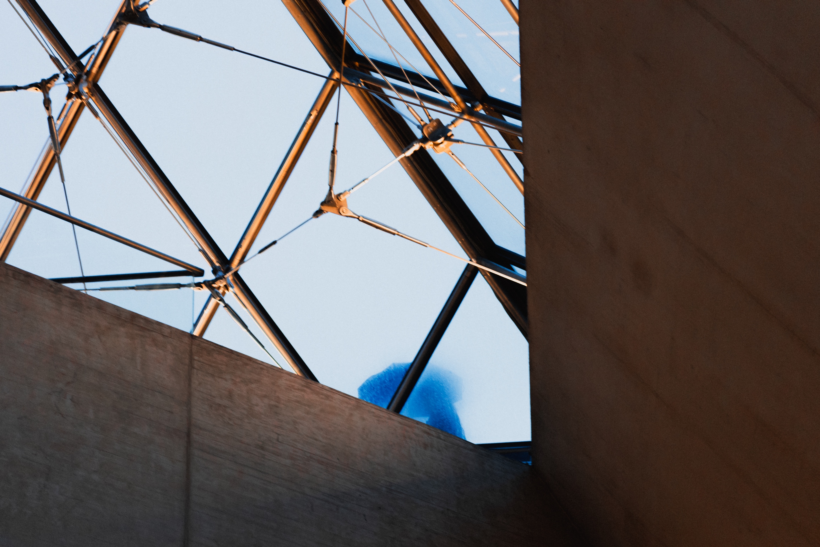 This image is a fascinating play of geometric patterns and light. The intersecting metal framework, highlighted by warm light, contrasts with the muted concrete and the cool tones of the sky. The triangular forms created by the structure draw the eye upward, adding depth and a sense of architectural dynamism. The composition is minimalist but striking, and the hint of a blue figure or object in the background adds an element of intrigue, breaking the rigid geometry with a softer, organic touch.