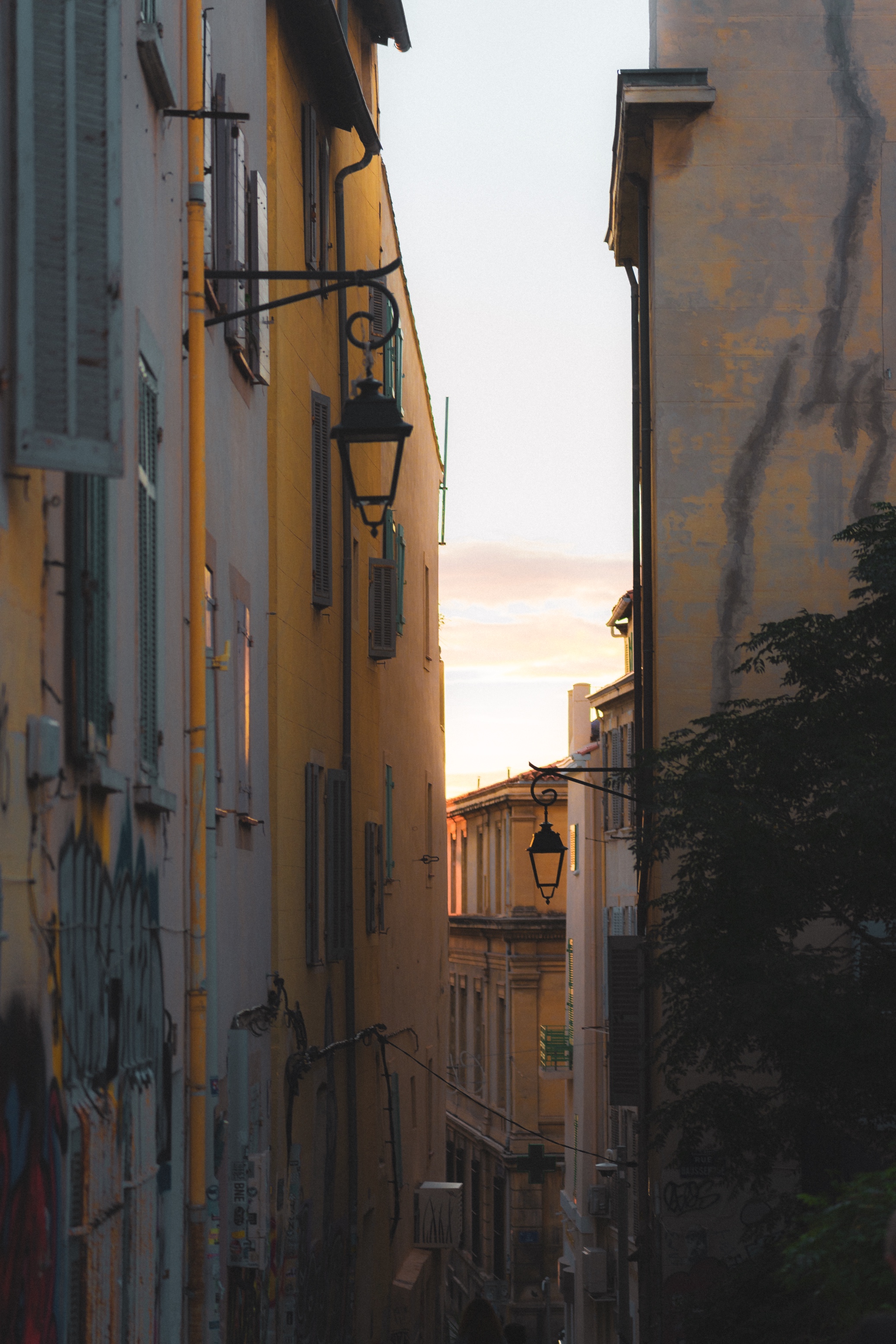 This photograph captures the essence of a quiet, narrow alley bathed in soft, golden light during what seems to be sunset or early evening. The warm tones of the buildings contrast beautifully with the cool green shutters and tree leaves, while the hanging lanterns add a nostalgic, old-world charm to the scene. The graffiti and weathered facades suggest an urban environment full of character and history. The way the light filters through the end of the alley draws the viewer’s attention deeper into the image, creating a sense of curiosity about what lies beyond.