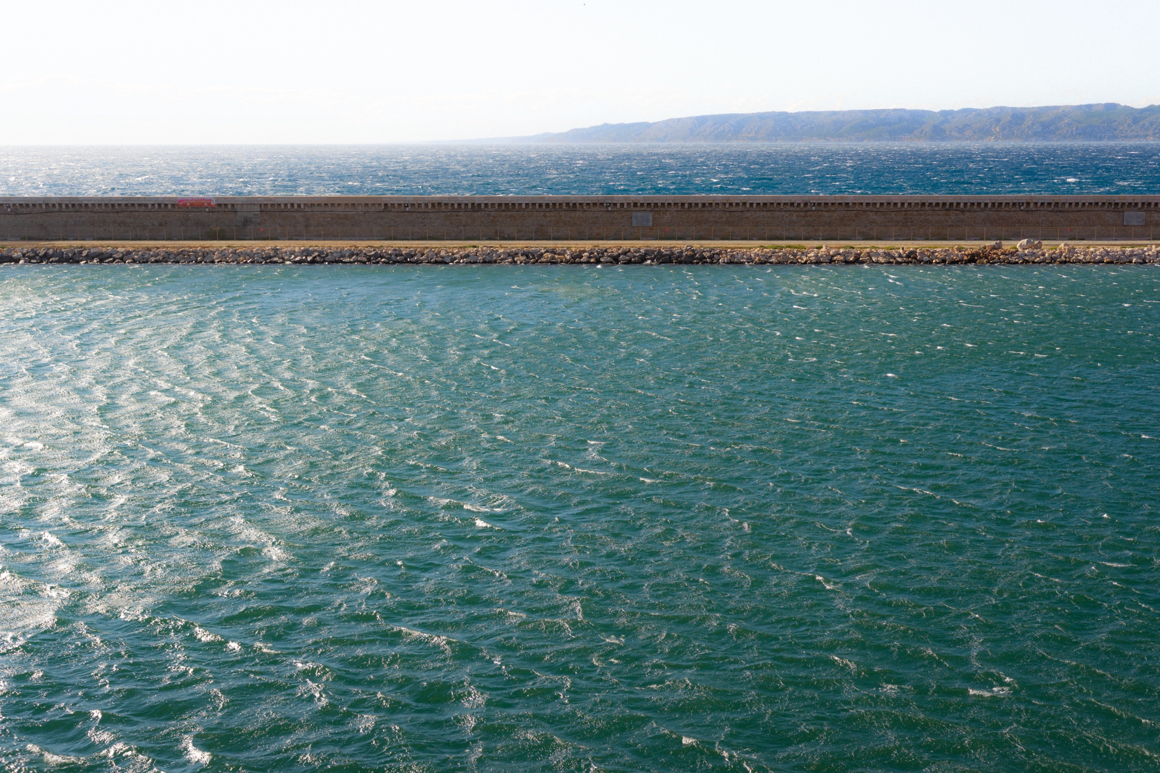 This image captures the dynamic interplay between the rippling water and the steadfastness of the barrier wall, with a distant mountain range providing depth to the composition. The texture of the water suggests a windy day, with sunlight glinting off the surface, adding an energetic vibrancy. The horizontal layering—the water, wall, and hills—creates a sense of balance and structure. The red train in the distance adds a pop of color and a human element to the otherwise natural and architectural scene.