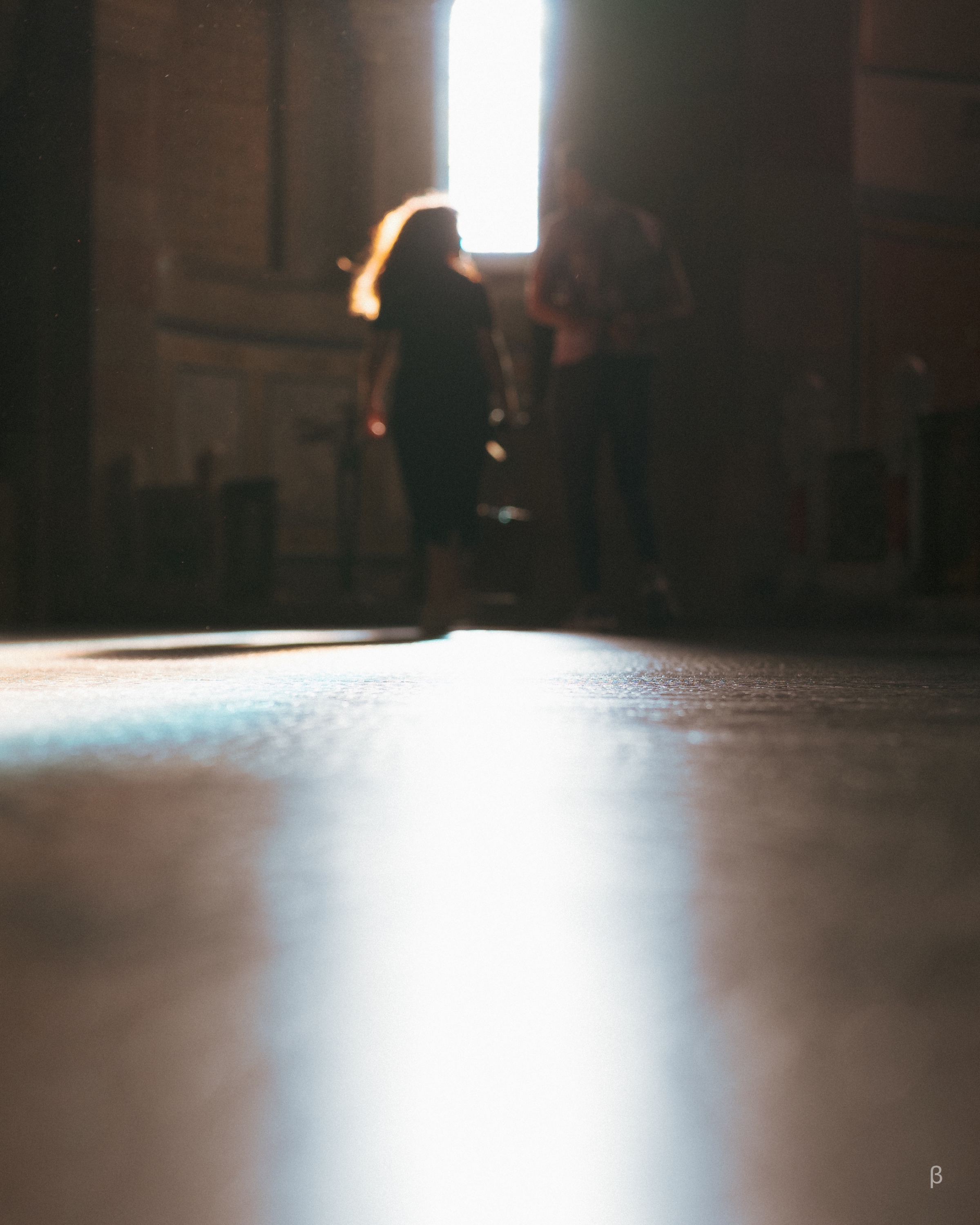 This image captures a wonderfully atmospheric moment, where two silhouetted figures stand near a bright window, their outlines softened by the intense backlight streaming in. The perspective from a low angle emphasizes the light reflecting off the floor, creating a dramatic contrast between the illuminated surface and the darker surrounding space. The glowing reflection leads the viewer’s eye toward the figures, adding depth and a sense of mystery to the scene. The overall mood feels ethereal and contemplative, suggesting themes like nostalgia, quiet encounters, or the passage of time in a serene and intimate environment. The warm tones and play of light evoke an emotional, almost cinematic quality.