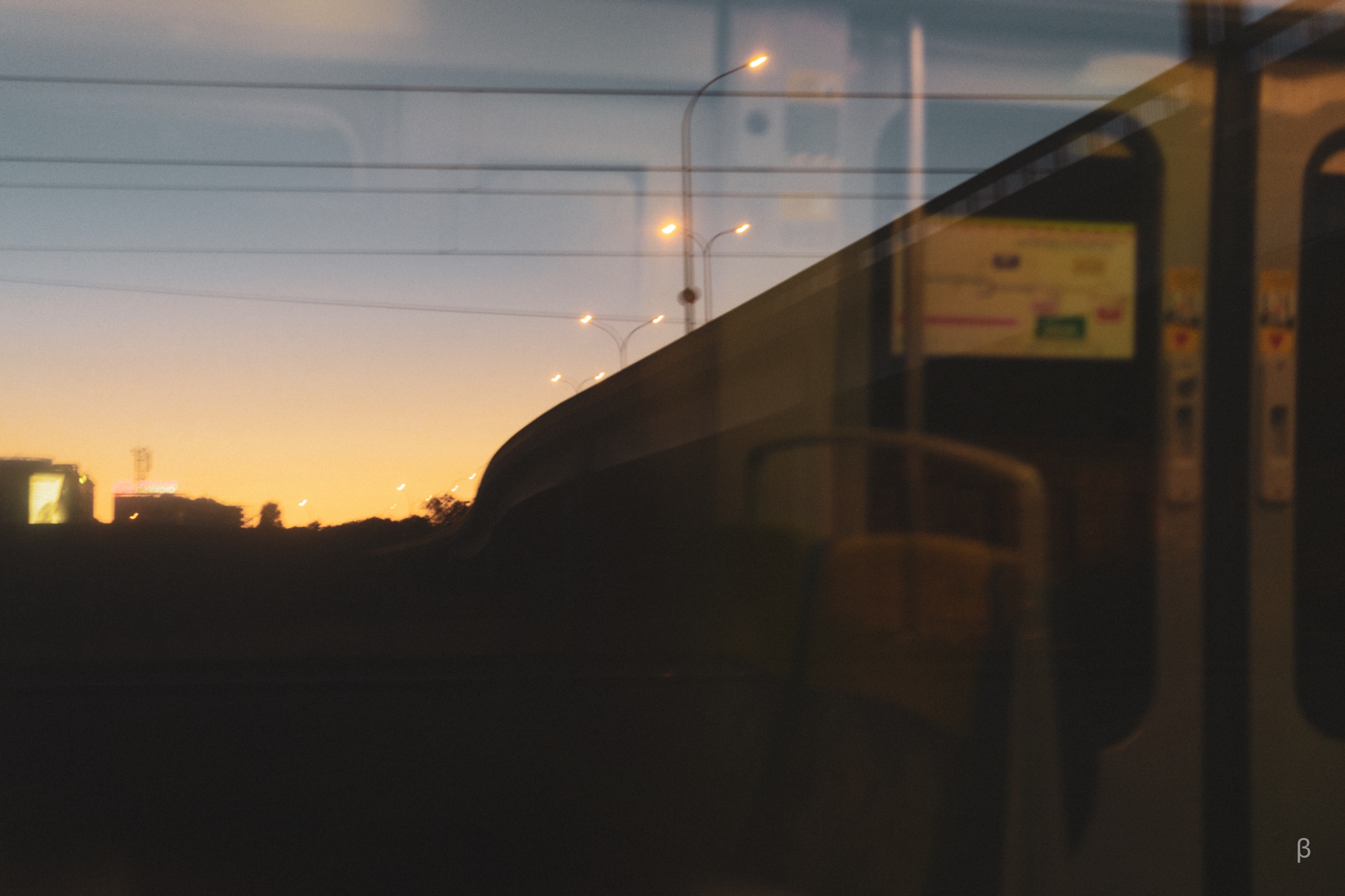 This image beautifully captures a layered perspective through a reflective surface, likely the window of a train or bus during sunset. The golden-orange glow of the evening sky illuminates the horizon, creating a peaceful contrast with the urban elements like lampposts, power lines, and silhouetted buildings outside. The reflection of the vehicle’s interior blends seamlessly with the outside view, merging rows of seats and signage into the scenery. This composition evokes a sense of motion and introspection, perfectly portraying the quiet, contemplative moments of a journey during twilight.