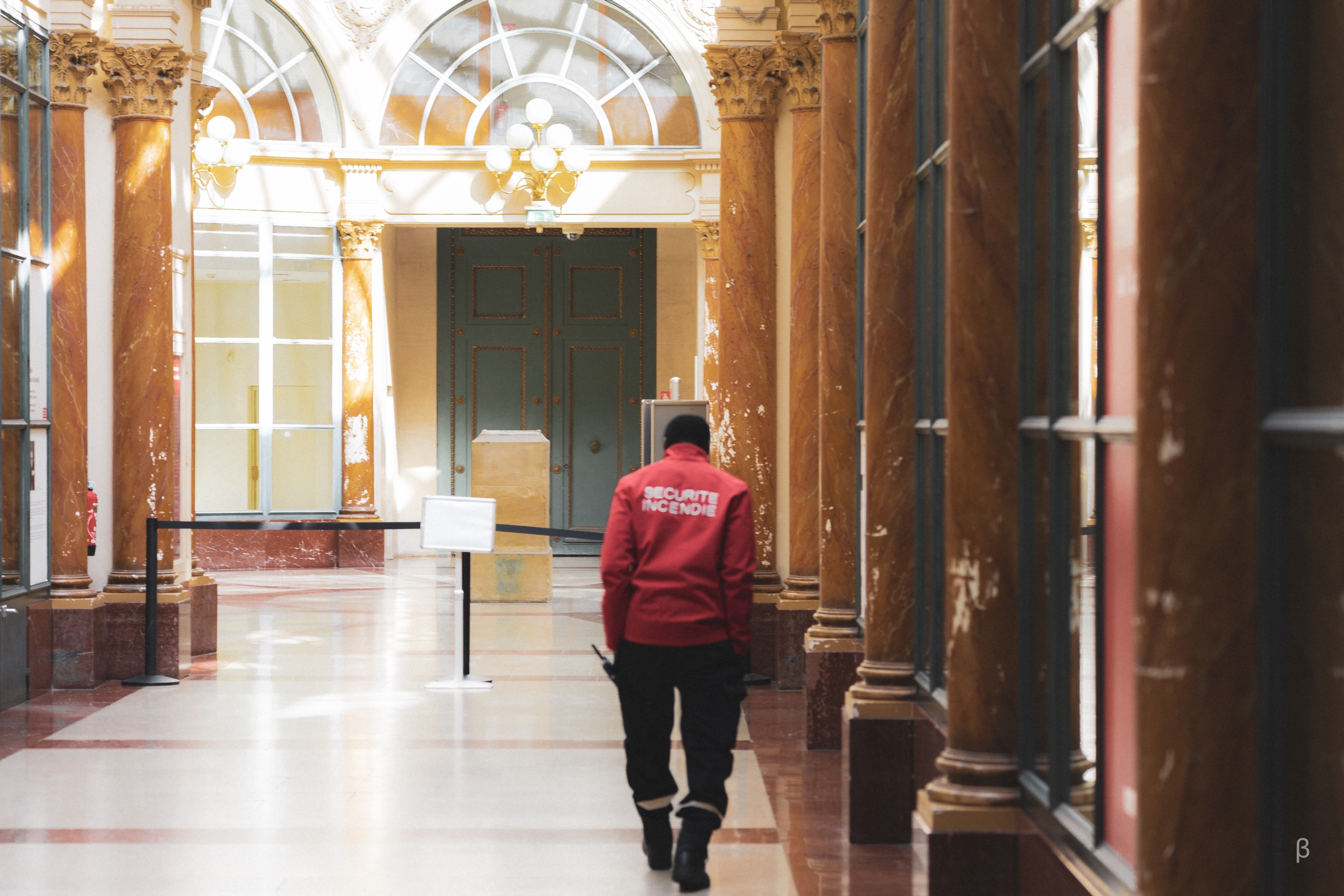 This photograph portrays a richly detailed interior space with a sense of historic elegance. Tall marble-like columns with intricate capitals frame the scene, while the light streaming through large, arched windows highlights the warm tones of the architecture. In the foreground, a security officer dressed in a red jacket with “Sécurité Incendie” written on the back walks away, adding a human element and a sense of purpose to the stillness of the space. The polished floors reflect the ambient light, creating a serene yet dynamic composition that emphasizes both structure and movement.