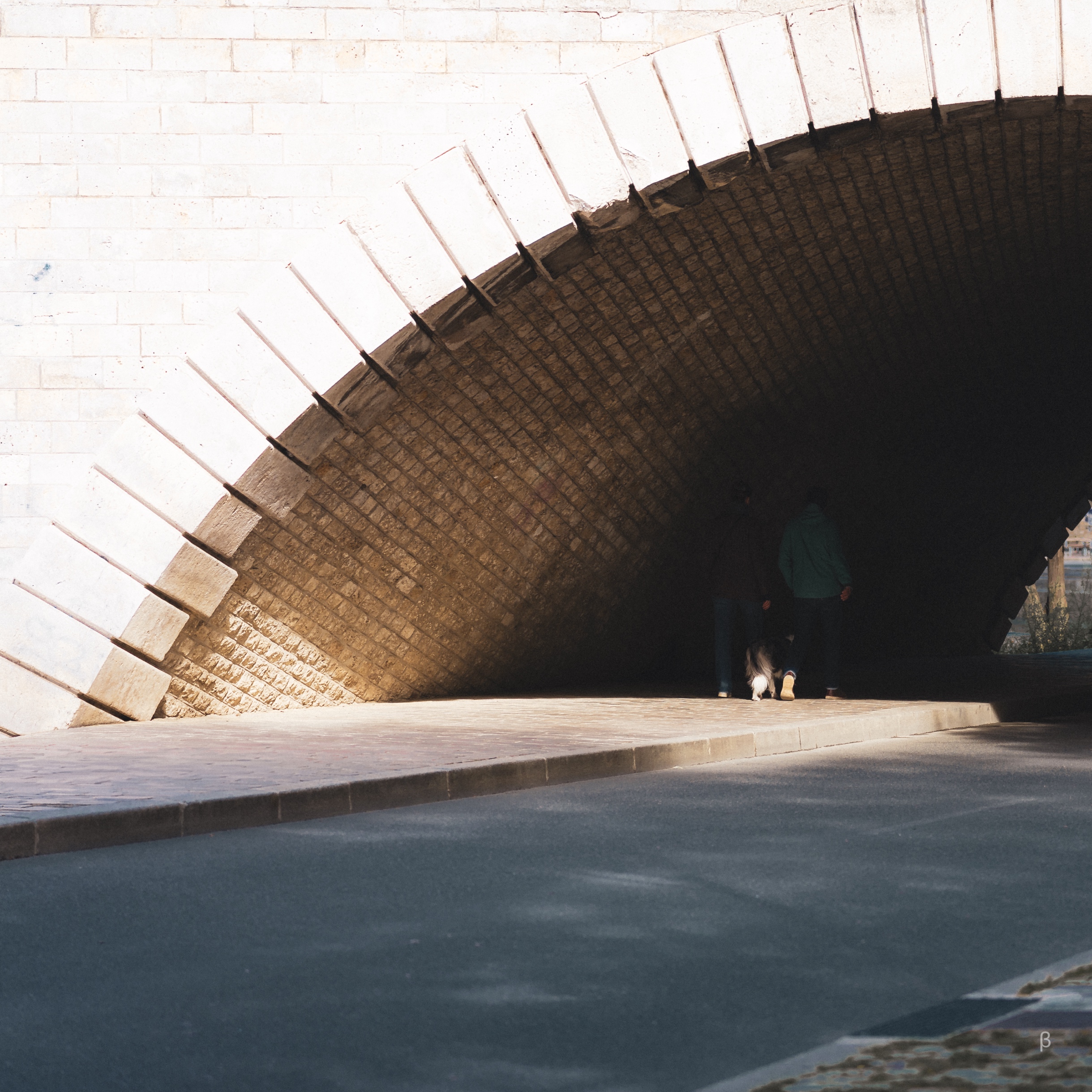 This photograph captures a serene urban moment beneath a beautifully arched stone bridge. The textured brickwork of the archway is illuminated by soft sunlight, creating a striking interplay of light and shadow. The scene is punctuated by two figures walking into the shadowed area, accompanied by a dog, adding a sense of quiet intimacy and movement to the otherwise tranquil setting. The foreground features a smooth, paved road, contrasting the rugged texture of the archway, while the soft tones and lighting evoke a peaceful, contemplative mood.