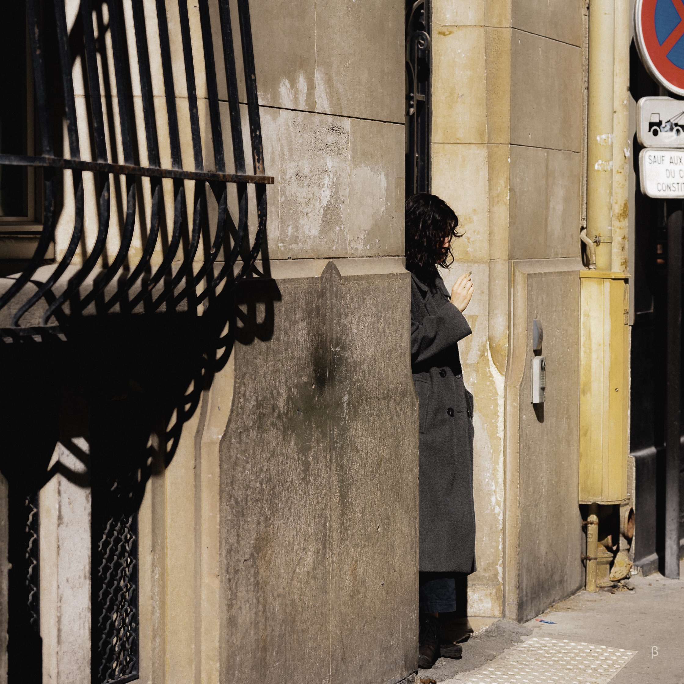 The image shows a person, likely a woman, standing alone in a dimly lit urban alley or passageway. The background is dominated by concrete walls, metallic grills, and other architectural elements that create a sense of seclusion and isolation. The person appears to be leaning against the wall, with their head down and shoulders hunched, suggesting a pensive or contemplative mood. The overall composition and lighting create a somber, melancholic atmosphere.