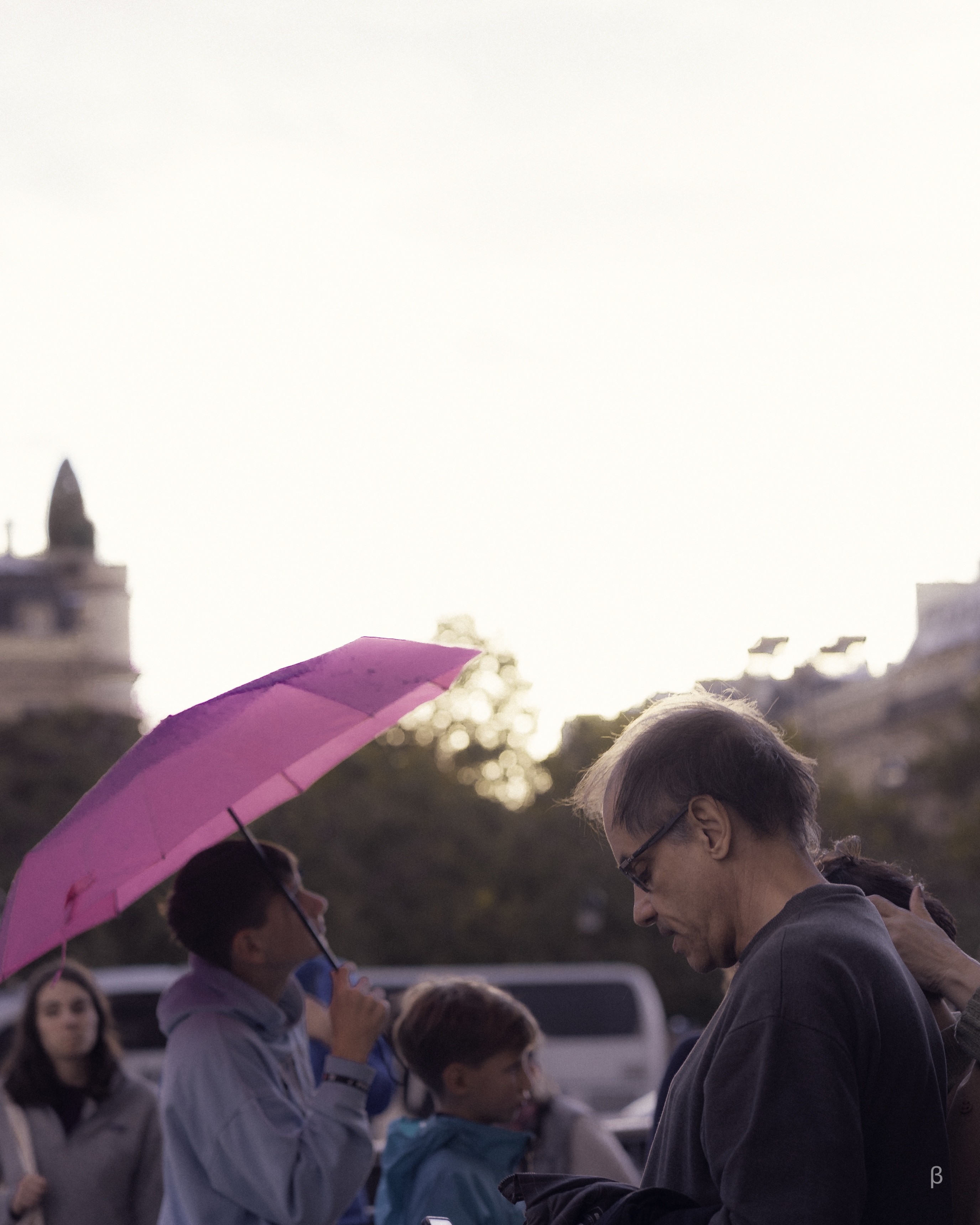 The image shows a group of people gathered together in an urban setting, with a person holding a pink umbrella in the foreground. The buildings and structures in the background appear to be blurred, creating a sense of focus on the people in the image. The people seem to be interacting and engaging with one another, though the specific details of their interactions are not clearly visible. The overall impression is one of a group of people seeking shelter or gathering together in a public space.