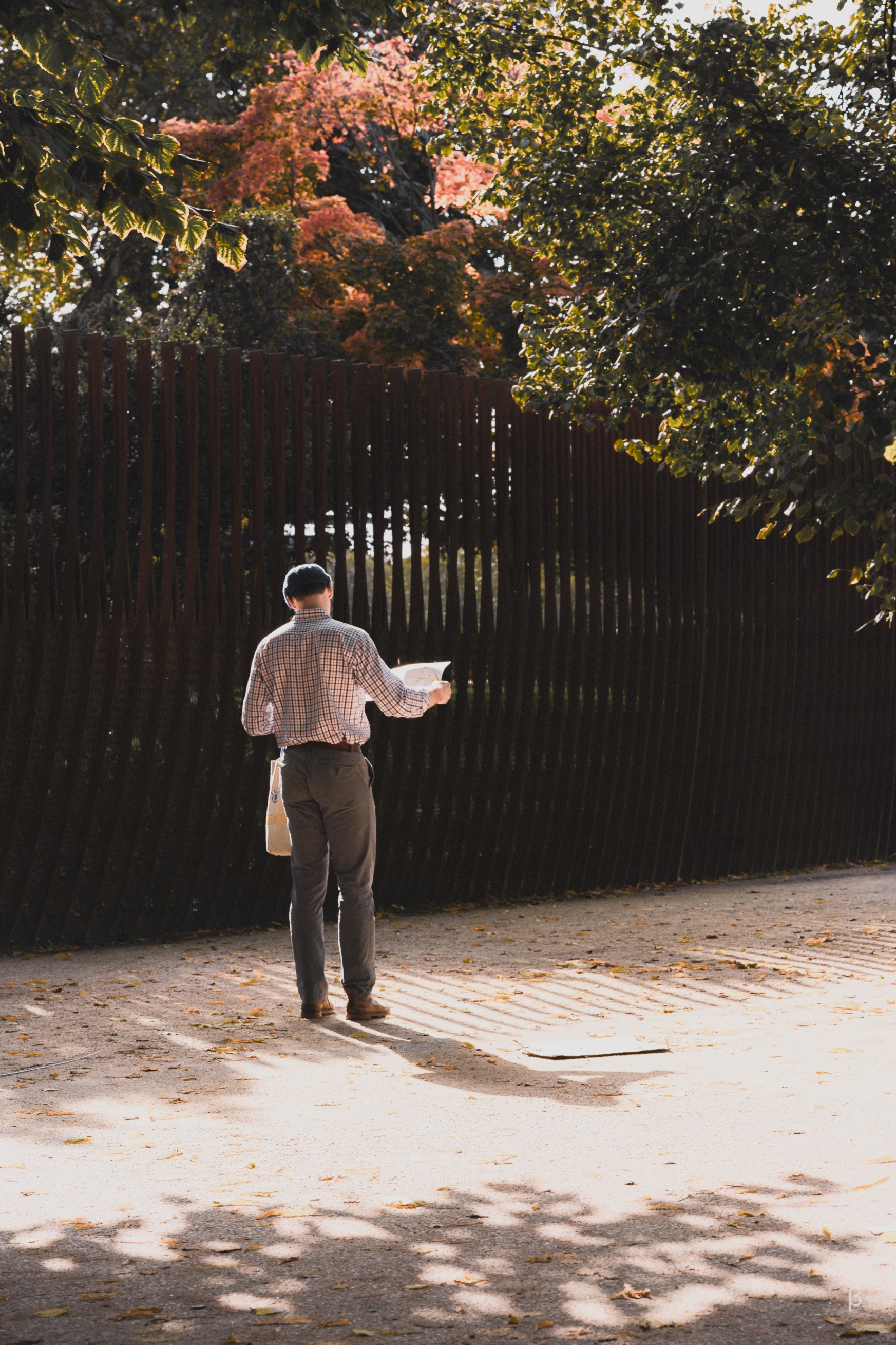 The image shows a man walking alone down a path in an autumn setting. The path is surrounded by a wooden fence, and the background has colorful fall foliage, with reds, oranges, and yellows visible in the trees. The man appears to be holding something, likely a book or paper, in his hands as he walks. The overall scene conveys a sense of solitude and contemplation in a peaceful, autumnal environment.