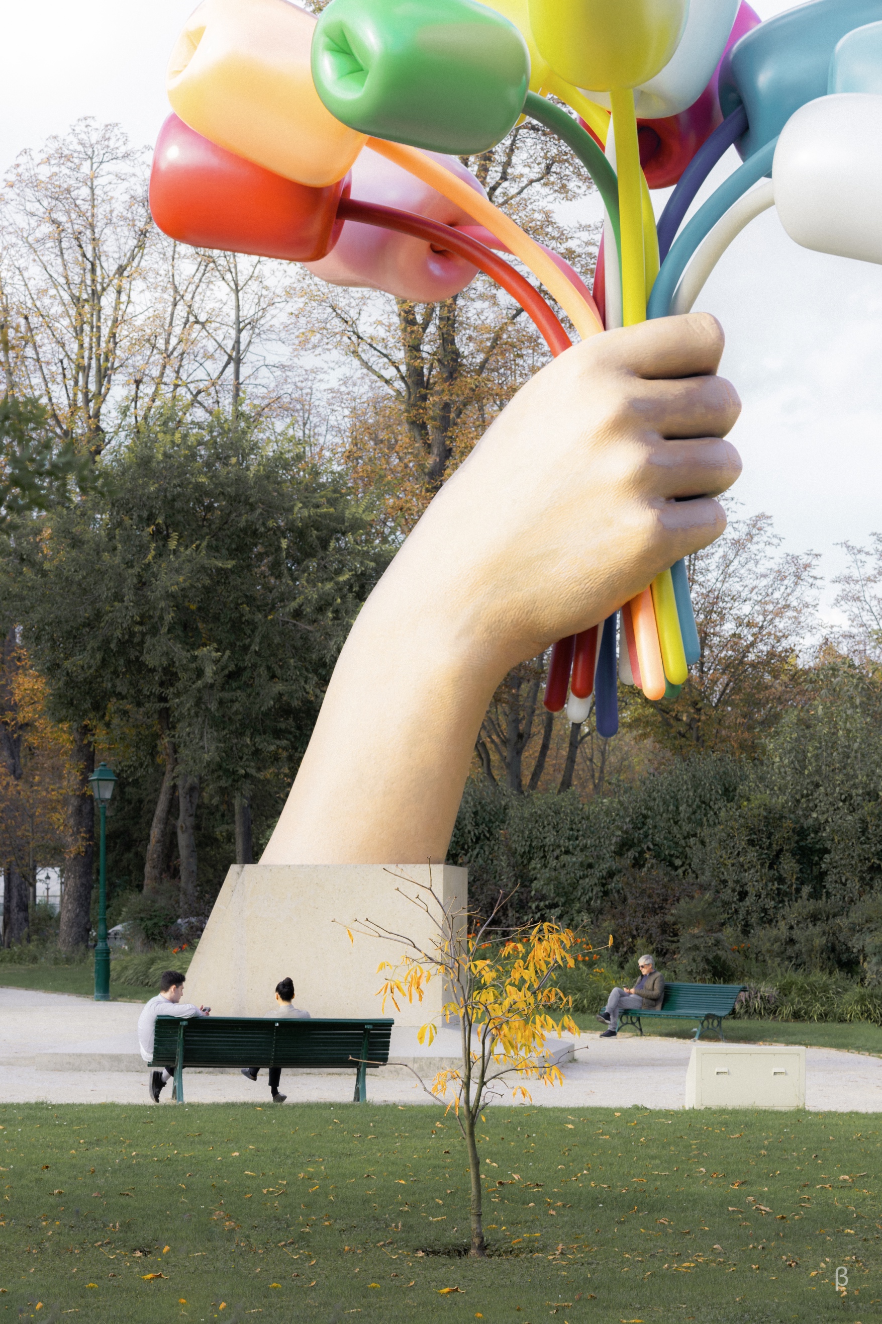 The image shows a colorful, abstract sculpture in the foreground, composed of large, curved, brightly colored tubes or shapes. In the background, there is a park-like setting with fall foliage, benches, and people sitting on them. The sculpture appears to be reaching up and over the space, creating an eye-catching and whimsical contrast with the natural surroundings. The combination of the bold, modern sculpture and the serene autumn scene makes for an interesting and engaging visual composition.