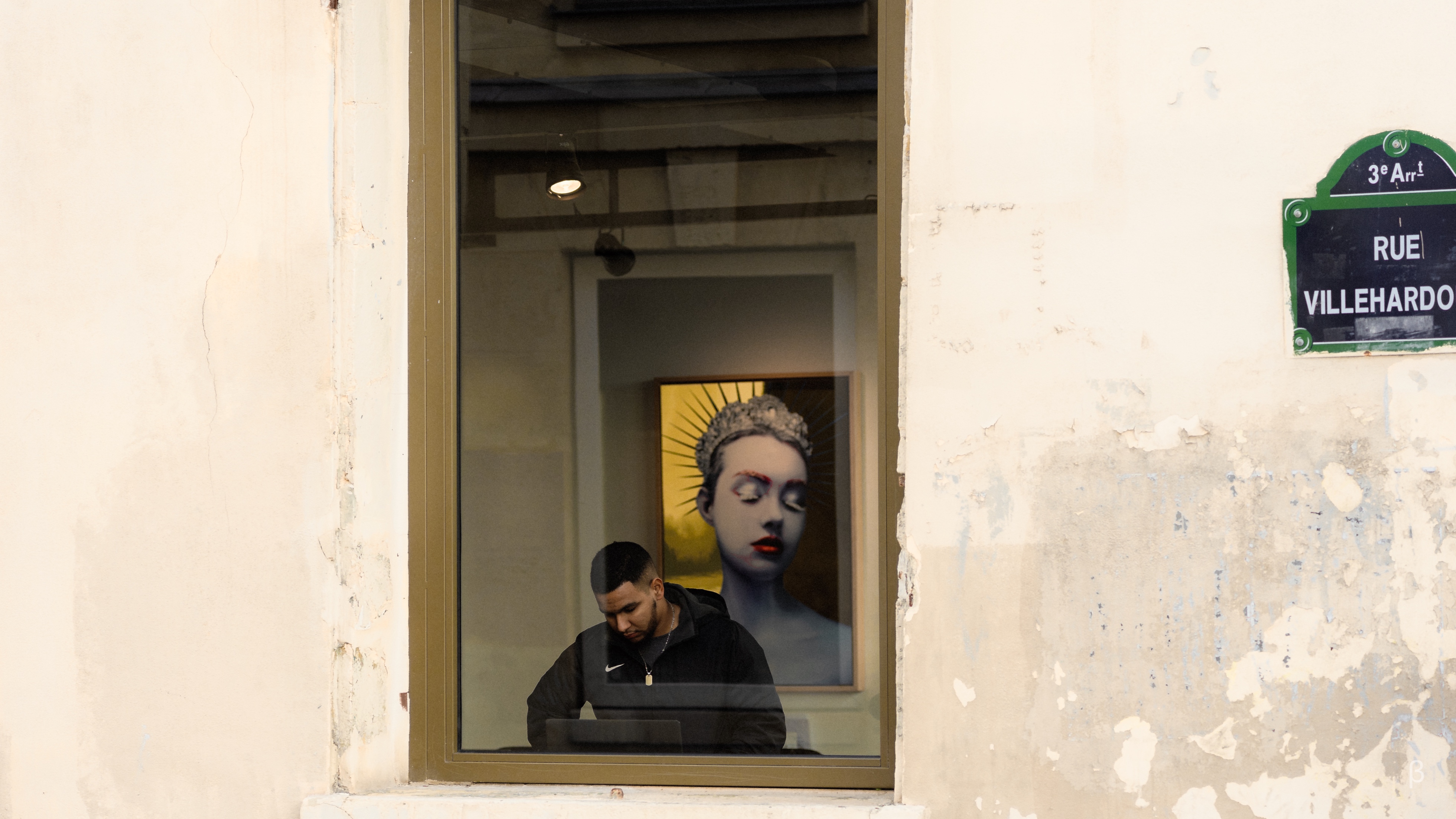 The image shows a man standing in the doorway of a building. Behind the man, through the window, there is a large sculptural figure with a distinctive headdress or crown. The background includes signage for &quot;Rue Villehardo&quot;. The man appears to be looking down, perhaps at his phone or other device. The contrast between the man in the foreground and the artistic figure in the background creates an interesting visual effect.