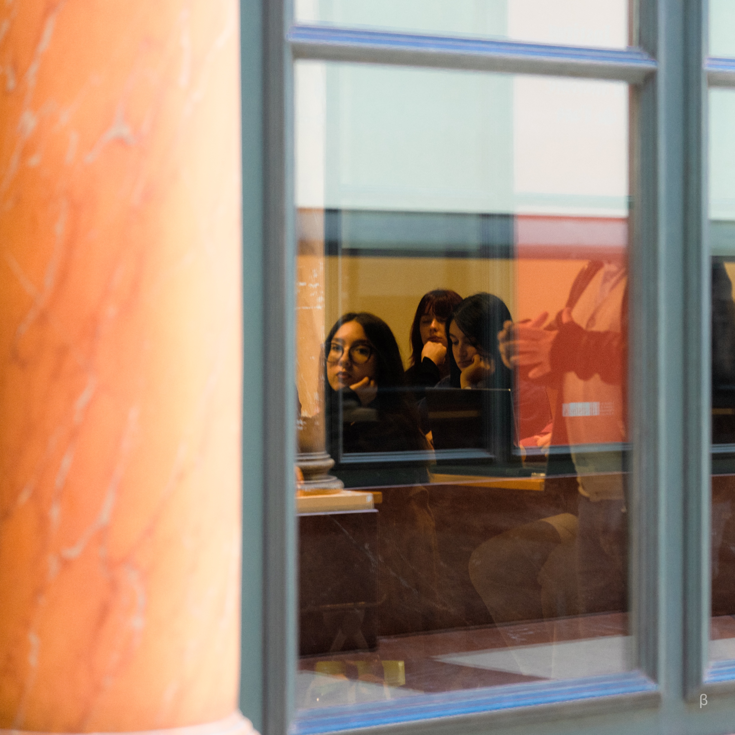 The image shows a reflection of three people visible through a window against an orange-tinted background. The reflection appears to be that of two women and a person sitting in a room or building, though their faces are not clearly visible. The window frame and the orange-colored wall or surface behind the window create an interesting visual effect, adding a sense of depth and perspective to the image.