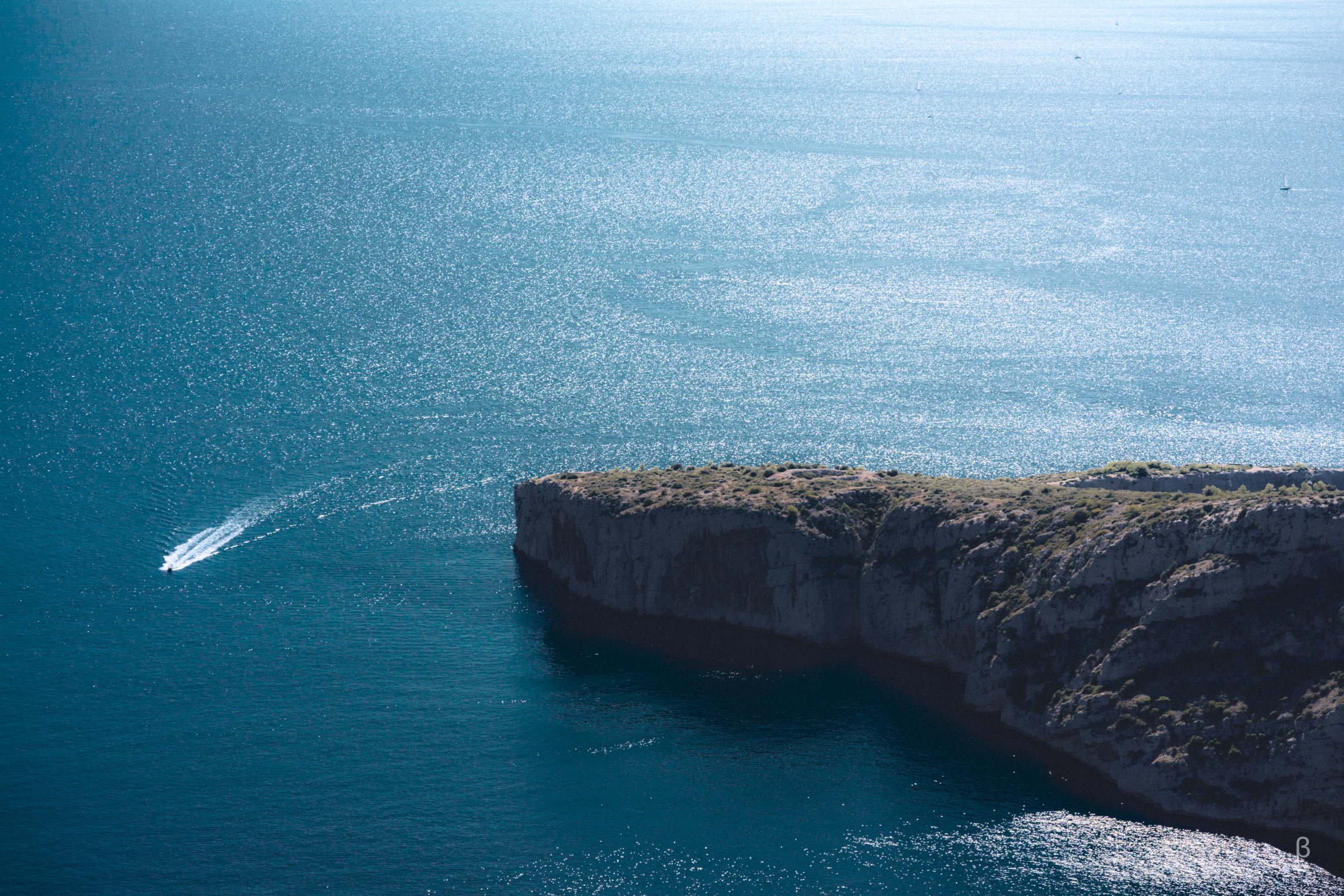 The image depicts a dramatic coastal landscape with a steep, rocky cliff overlooking a vibrant turquoise sea. A small boat is visible speeding across the water in the distance. The overall scene conveys a sense of rugged, natural beauty.