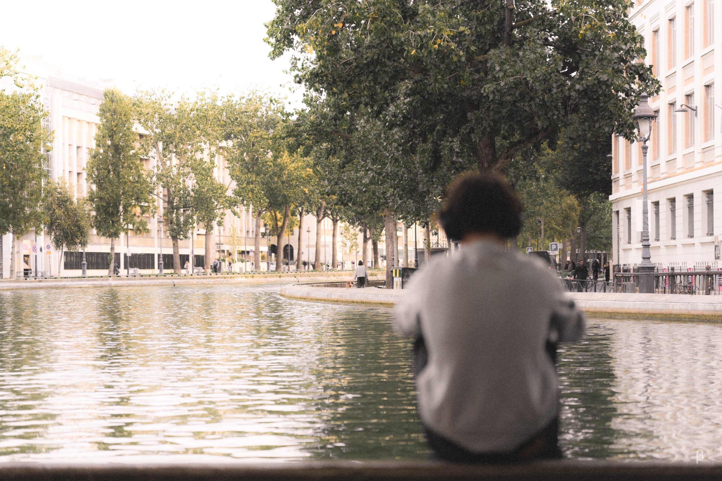 The image shows a tranquil urban plaza with a central water feature. Tall, mature trees line the perimeter, casting dappled shade over the scene. A person with curly dark hair is seated on the edge of the water, gazing outward. The overall atmosphere conveys a sense of calm and relaxation in a well-designed public space.