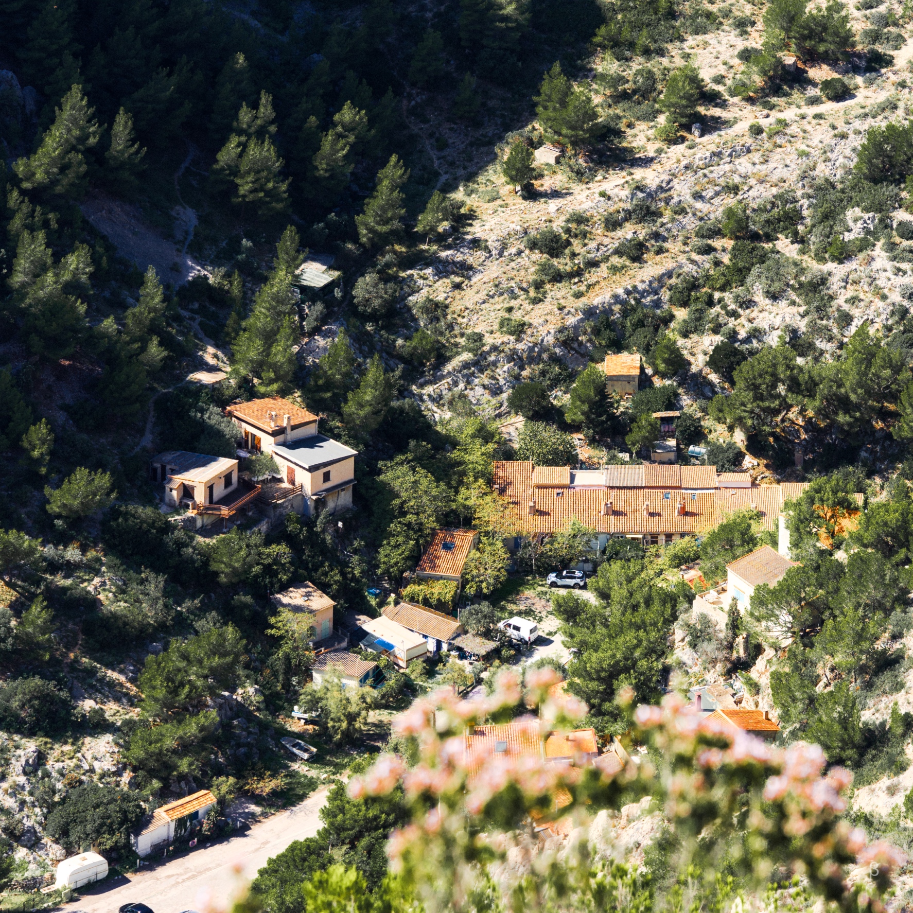 The image shows a mountainous, forested landscape with several small buildings nestled among the trees. The buildings appear to be houses or cabins, with reddish-orange tiled roofs that contrast against the lush green foliage. The terrain is steep and rocky, with a mix of pine and other evergreen trees covering the slopes. The overall scene conveys a remote, rustic setting in a scenic, mountainous area.