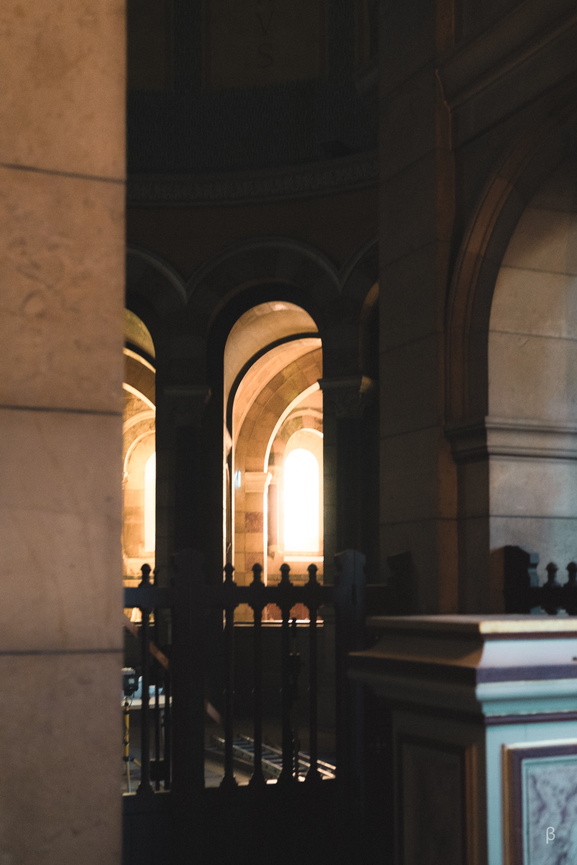 The image shows a dimly lit interior space with a series of arched doorways or windows. The archways are backlit, creating a dramatic contrast between the bright, warm light in the distance and the shadowed foreground. The architecture appears to be of a classical or historic style, with decorative elements visible along the edges of the arches. The overall composition creates a sense of depth and mystery, drawing the viewer's eye through the layered frames towards the illuminated space beyond.