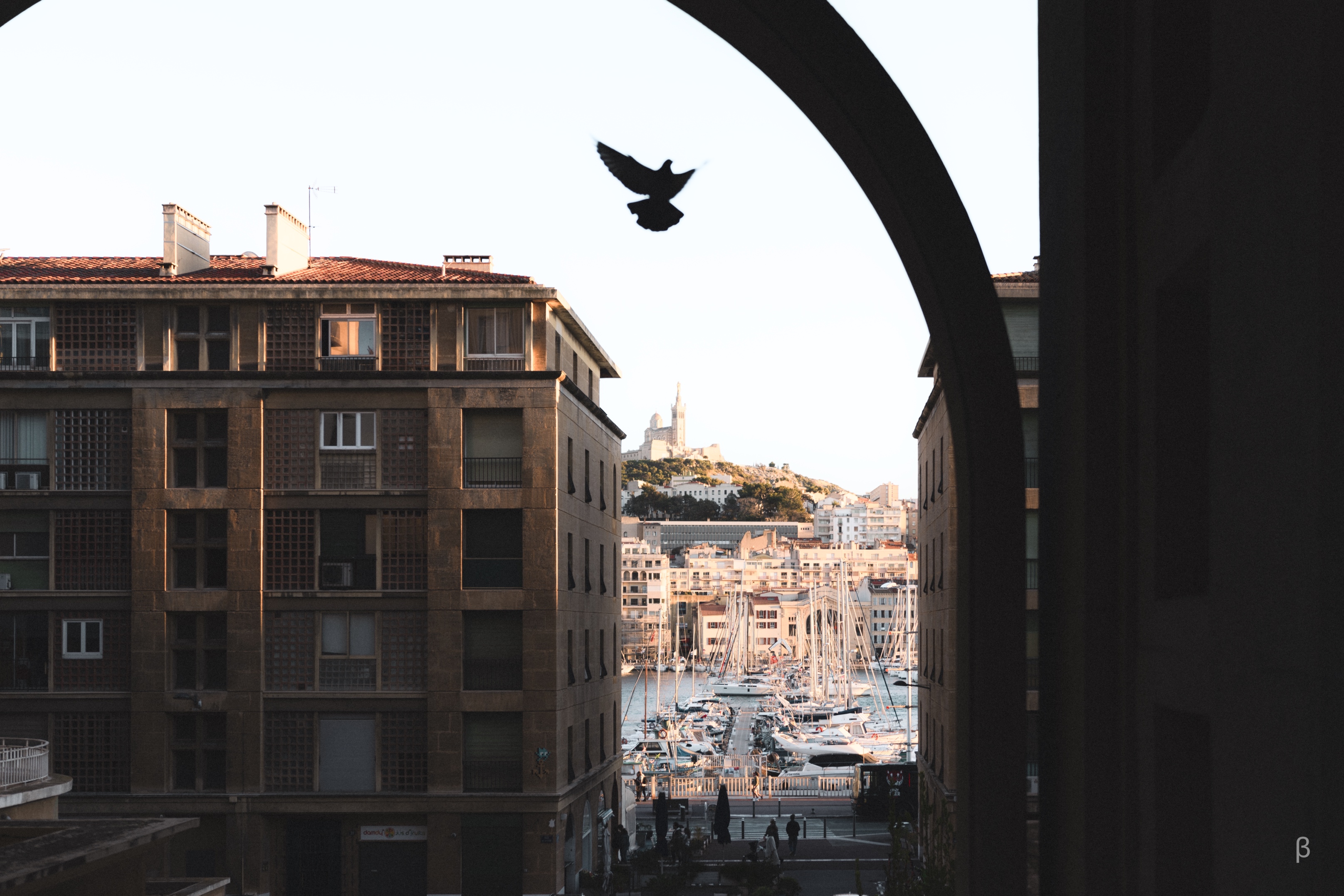 The image shows a scenic view of a harbor area in what appears to be a Mediterranean city. In the foreground, the frame is defined by the arched architectural structure, which creates a shadowed silhouette against the bright sky. Through this archway, the viewer can see the bustling harbor below, with rows of sailboats and yachts moored at the docks. In the distance, the cityscape rises up the hillside, with a distinctive church or tower visible at the top. The overall composition highlights the contrast between the historic, shadowed architecture in the foreground and the vibrant, sun-drenched harbor scene beyond.