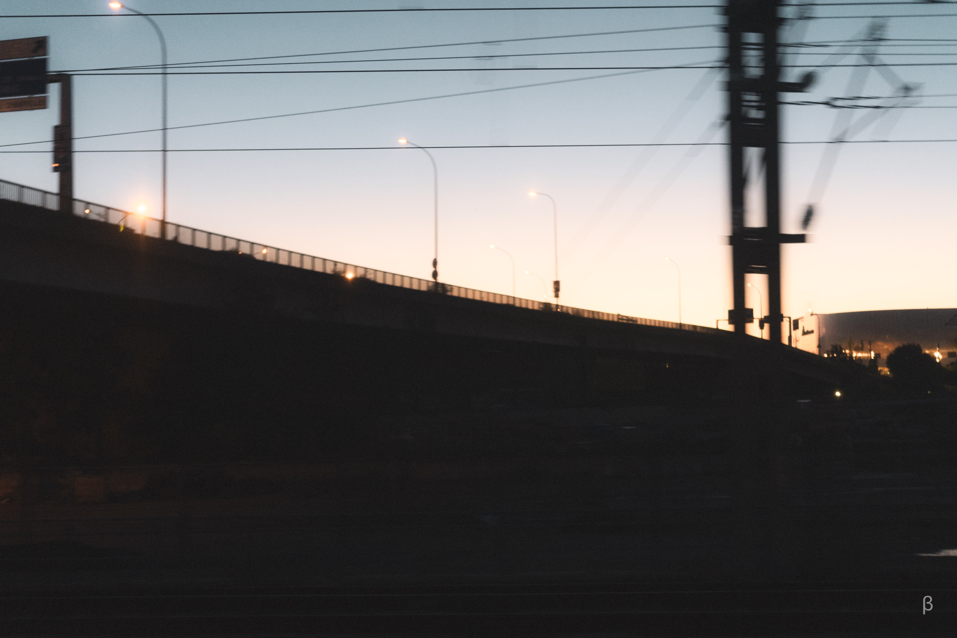 The image shows a nighttime view of a long bridge or overpass, with tall concrete or steel support structures and an expanse of road or rail tracks visible in the distance. The sky is hazy and bathed in a warm, pinkish-orange glow, suggesting a sunset or twilight scene. Streetlights and other illumination sources line the bridge, creating a sense of movement and energy in the frame. The overall composition creates an atmospheric, almost cinematic impression of urban infrastructure set against the backdrop of the changing sky.