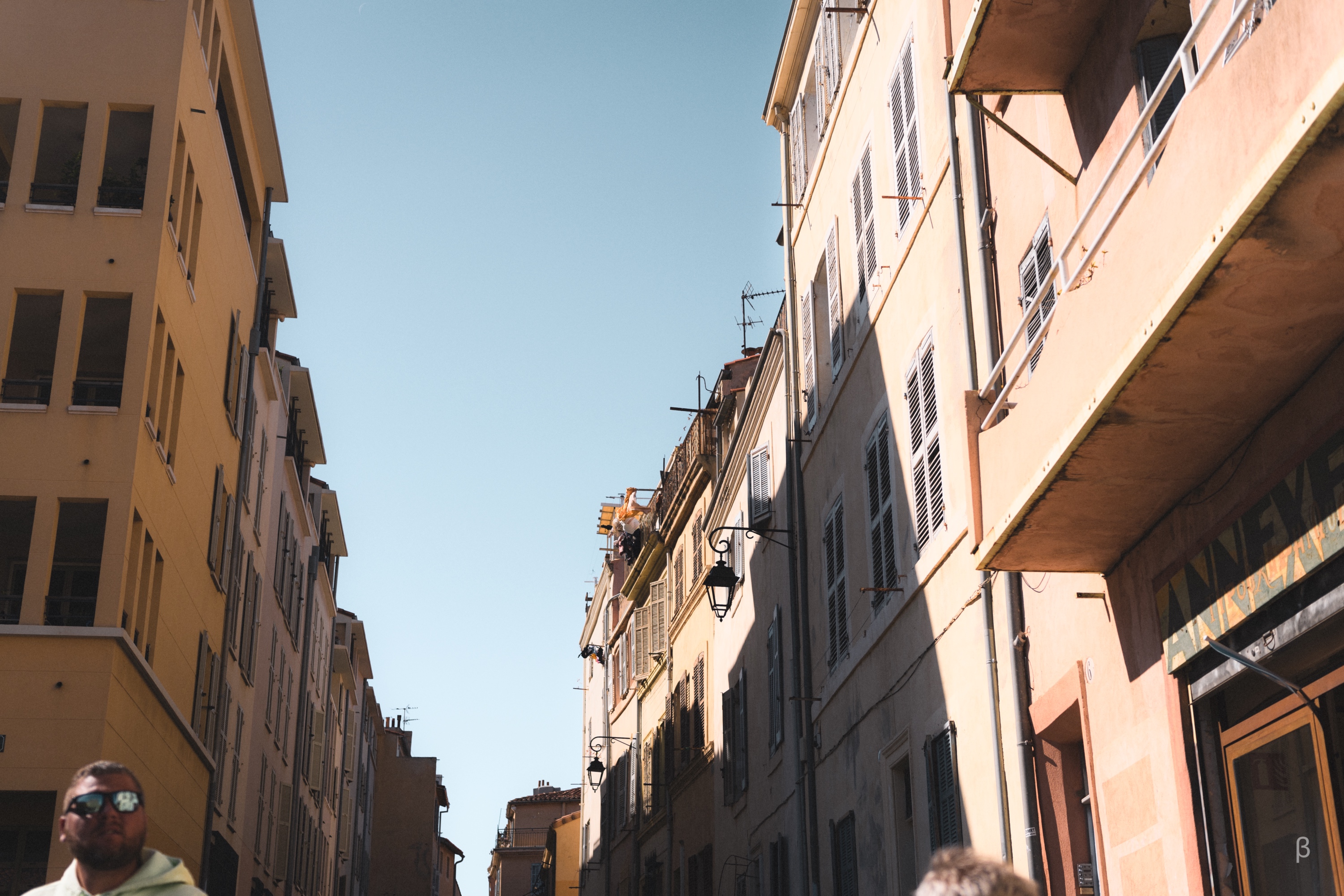 The image appears to show a narrow street in a European city, with rows of multi-story buildings lining the sides. The buildings have a warm, ochre-colored facade and feature various architectural elements like shuttered windows, balconies, and hanging wires. The sky is bright and clear, indicating a sunny day. The overall scene conveys a quaint, historic feel typical of Mediterranean urban settings.