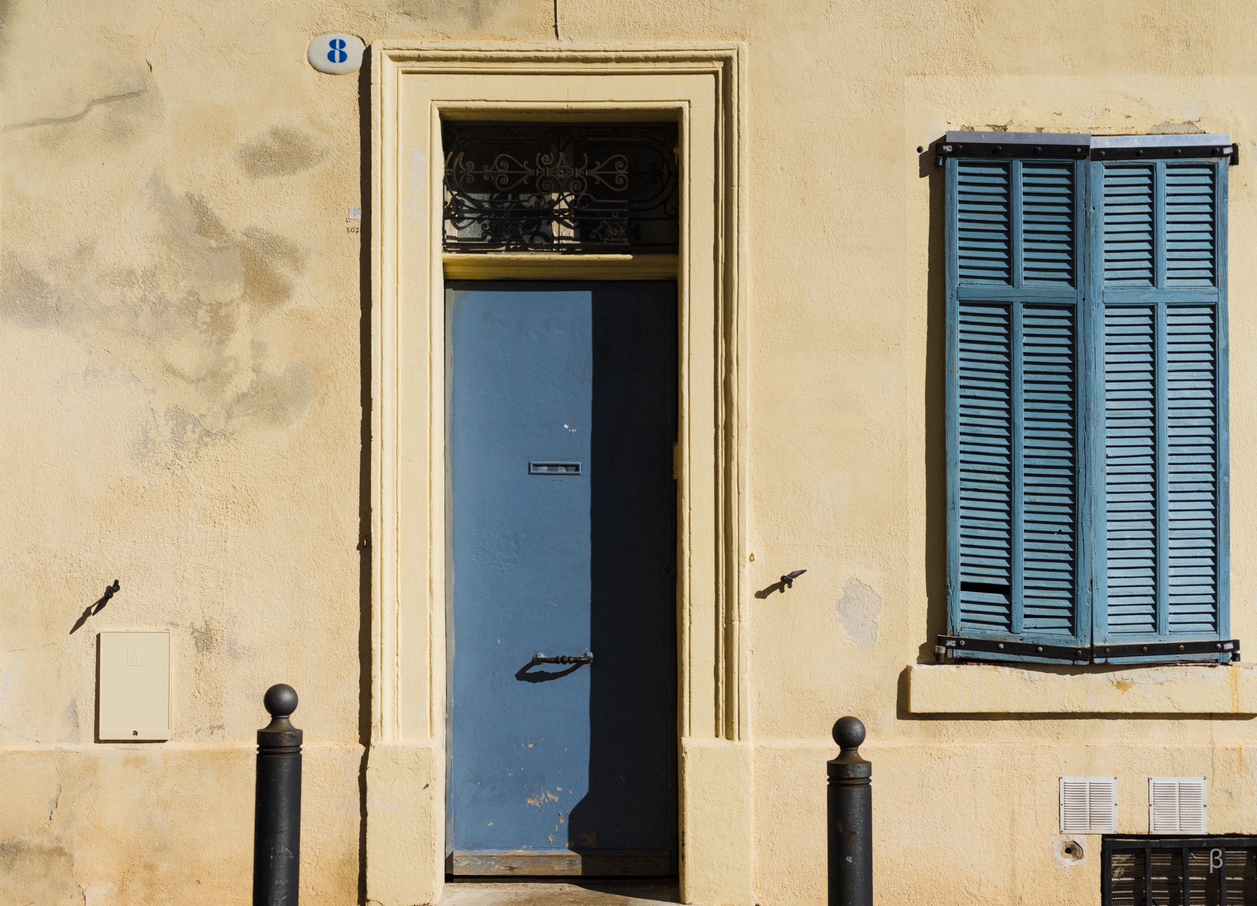 This image depicts a doorway set into a weathered, beige exterior wall. The doorway has an ornate, golden frame, and the door itself is a solid, dark blue color. Next to the doorway are blue louvered shutters on a window. There is also a black post or bollard visible near the entrance. The overall scene has an aged and textured appearance, with cracks and wear visible on the wall surface. The contrast between the warm beige wall and the cooler blue tones of the door and shutters creates an interesting visual dynamic.