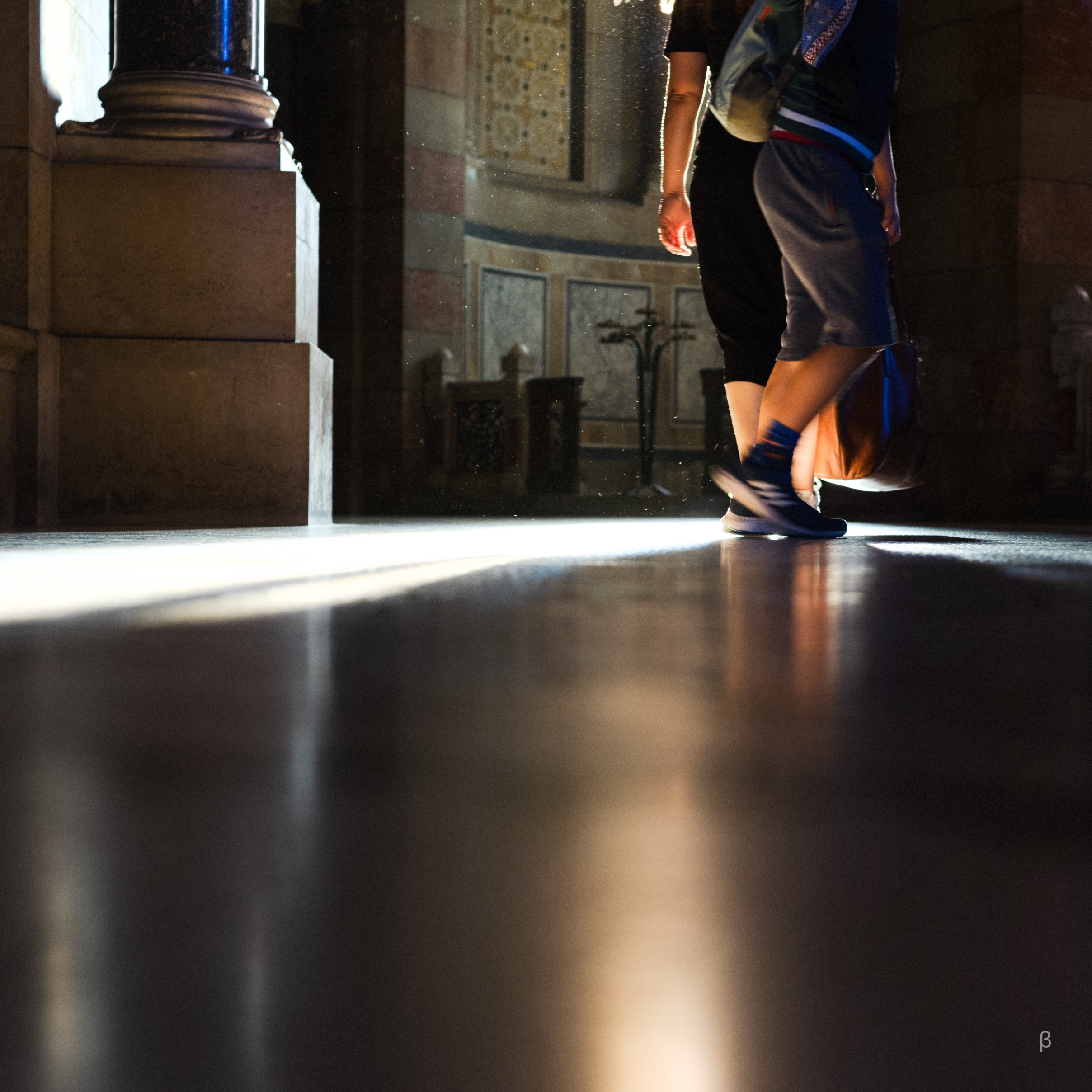 The image depicts a dimly lit hallway with strong contrasts between light and shadow. A silhouetted figure is visible at the end of the walkway, near a set of illuminated double doors. The composition creates a moody, atmospheric effect with the interplay of light and shadow.