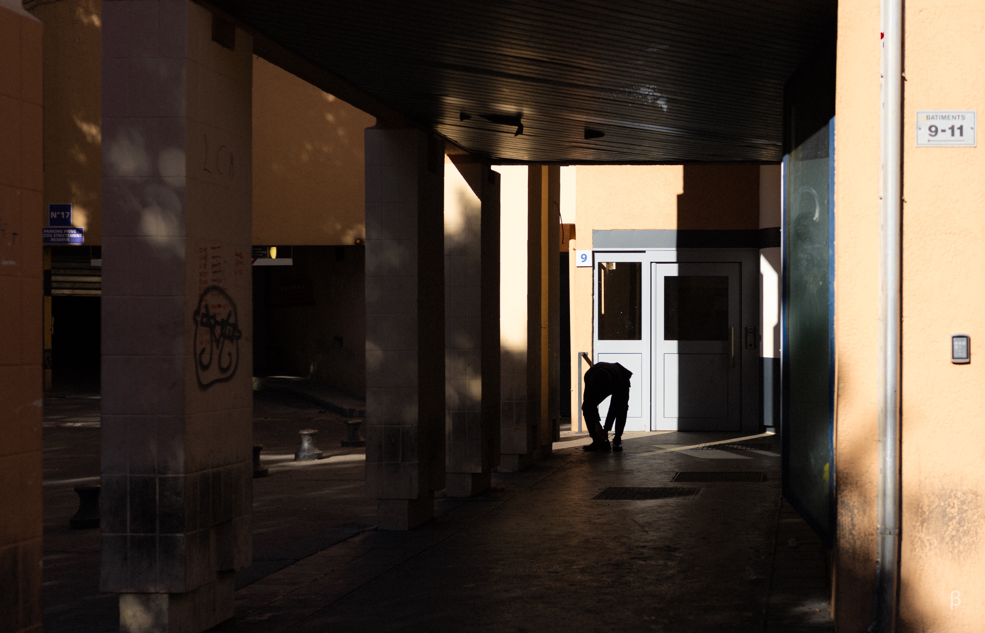 A dimly lit covered walkway or arcade with concrete pillars casting long shadows. At the end of the walkway is a white double door entrance illuminated by natural sunlight. A silhouetted figure is bent over near the doorway. The scene has strong contrast between dark shadows and warm sunlit areas. A building number '9-11' is visible on a sign, and there appears to be some graffiti on one of the pillars. The composition creates a moody, atmospheric effect through the interplay of light and shadow.