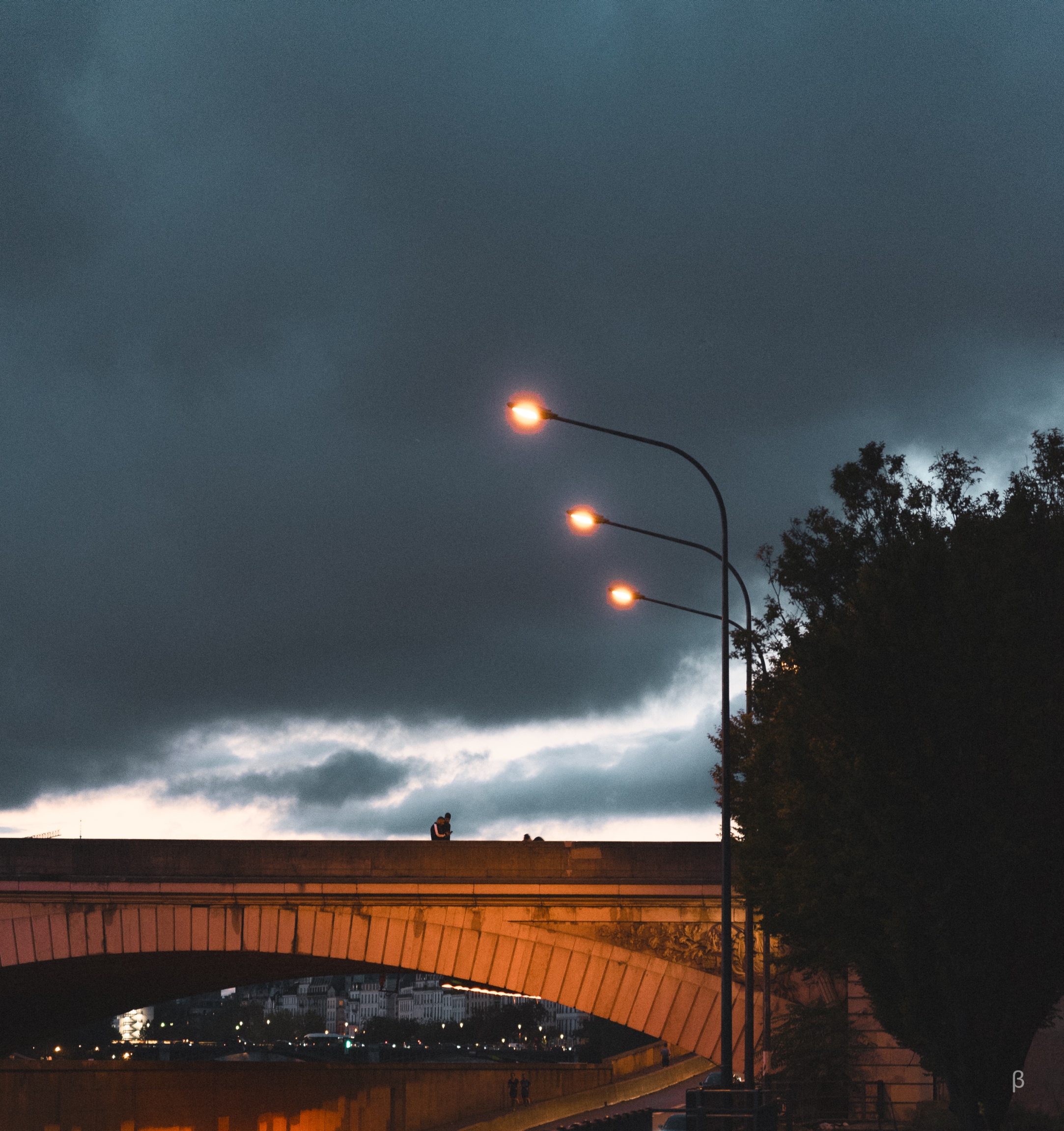 This image depicts a nighttime urban scene with dramatic clouds in the sky. A pedestrian bridge spans a body of water, illuminated by warm lighting. Several streetlights cast a glow over the area. The overall atmosphere feels moody and atmospheric.