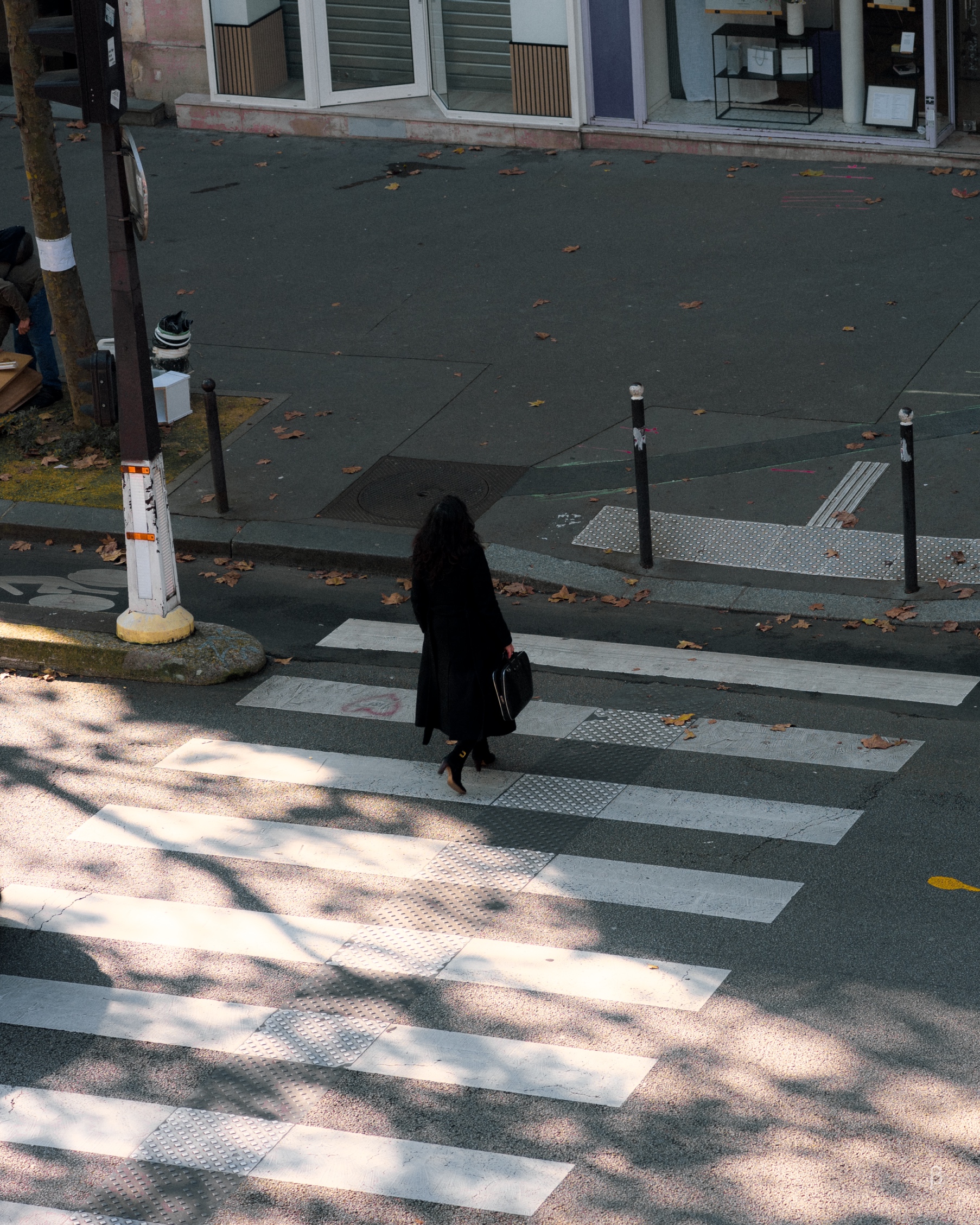 A person in a long black coat crosses a zebra crossing on an autumn day. Scattered fallen leaves dot the street, and dappled sunlight creates patterns across the white pedestrian stripes. The scene appears to be in an urban setting, with a storefront visible in the background.