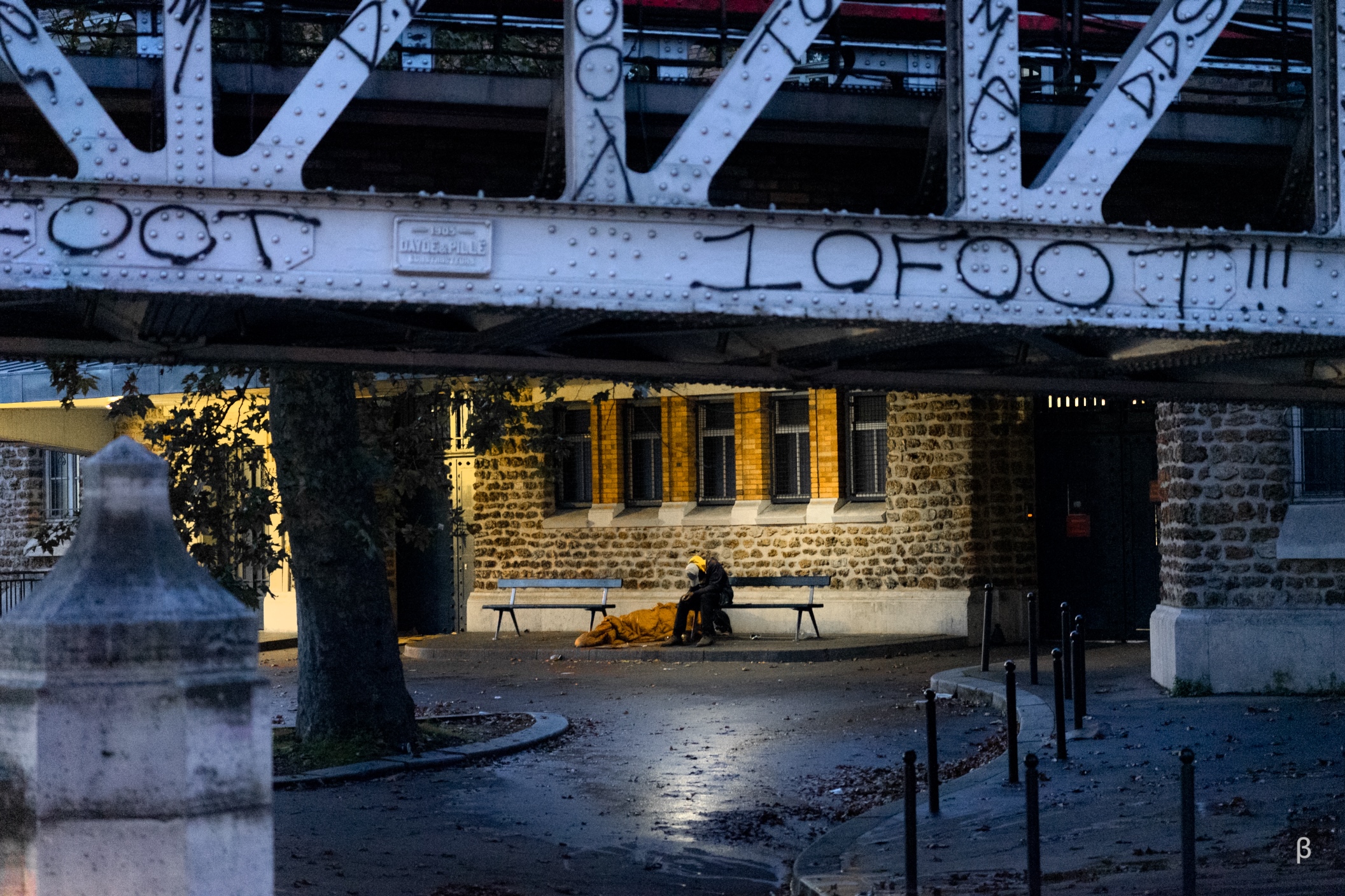 The image shows a nighttime urban scene under a large metal bridge structure. The bridge has intricate metalwork and the word &quot;LOFOT&quot; is visible on the side. Below the bridge, there is a stone building with illuminated windows, and a person sitting on a bench in the foreground. The scene has a moody, atmospheric quality with the mix of light and shadows.