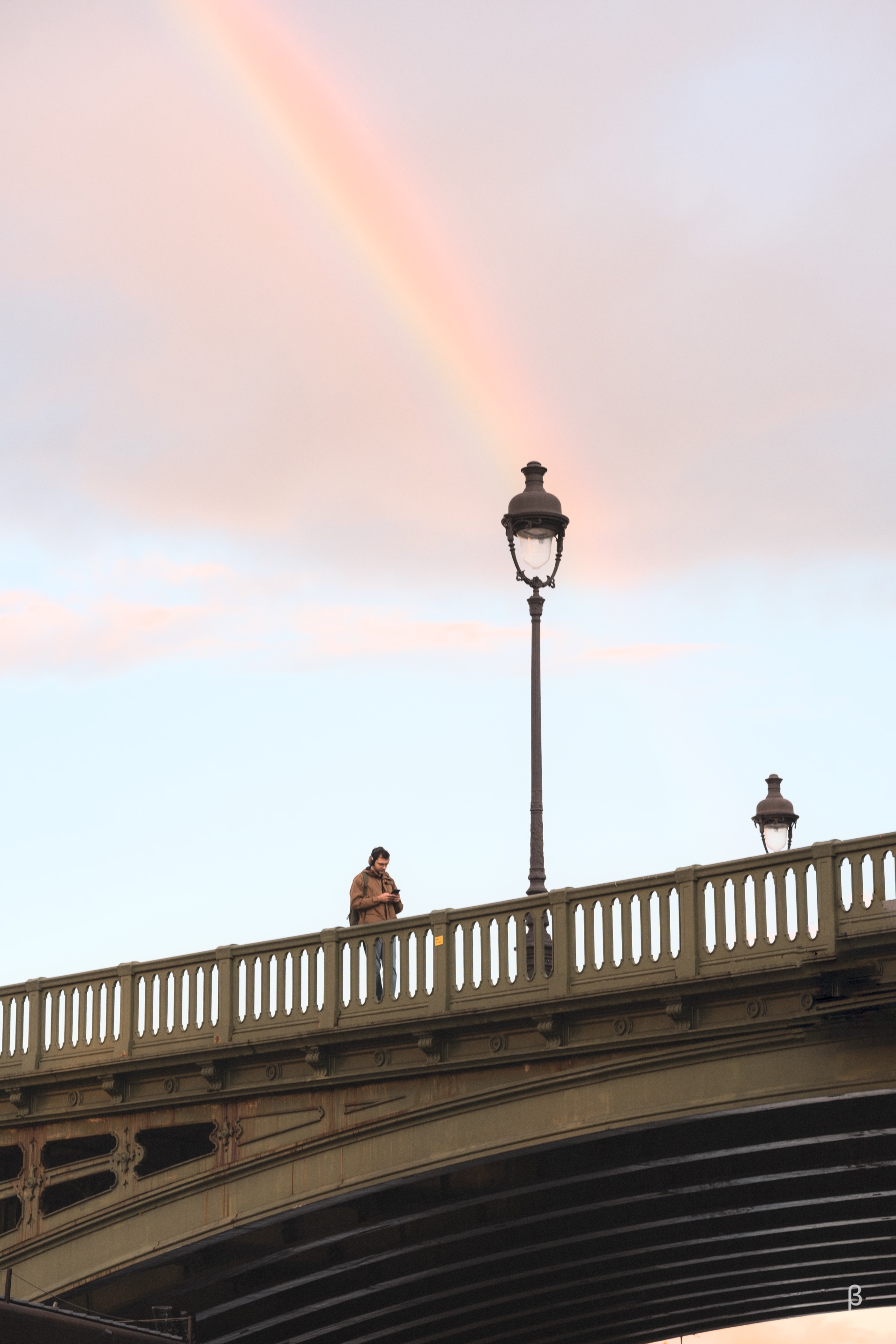 The image shows a person standing alone on a bridge, silhouetted against the dramatic, colorful sky at sunset. The bridge structure features ornate railings and architectural details, giving it a historic, European feel. The person is positioned in the center of the frame, creating a sense of contemplation or solitude within the urban landscape. The contrast between the warm, vibrant sky and the dark, shadowed figure on the bridge adds to the moody, atmospheric quality of the scene.