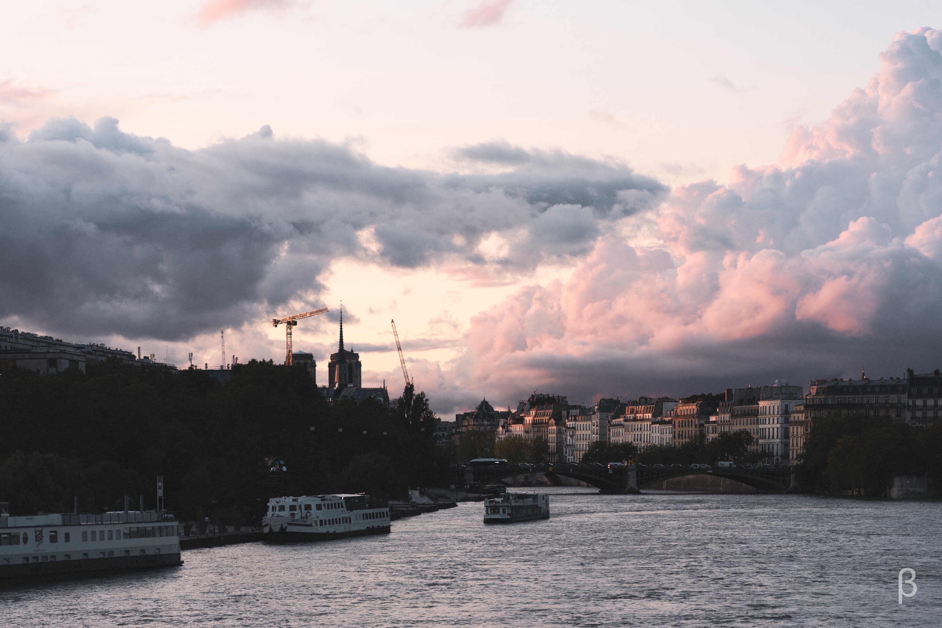 The image depicts a scenic view of Paris, France. It shows the Seine River flowing through the city, with the iconic Notre-Dame Cathedral visible in the distance, partially obscured by surrounding buildings and cranes. The sky is filled with dramatic, colorful clouds, creating a striking contrast between the dark, moody tones of the cityscape and the vibrant, warm hues of the sunset. The image captures the dynamic and picturesque nature of the Parisian landscape.