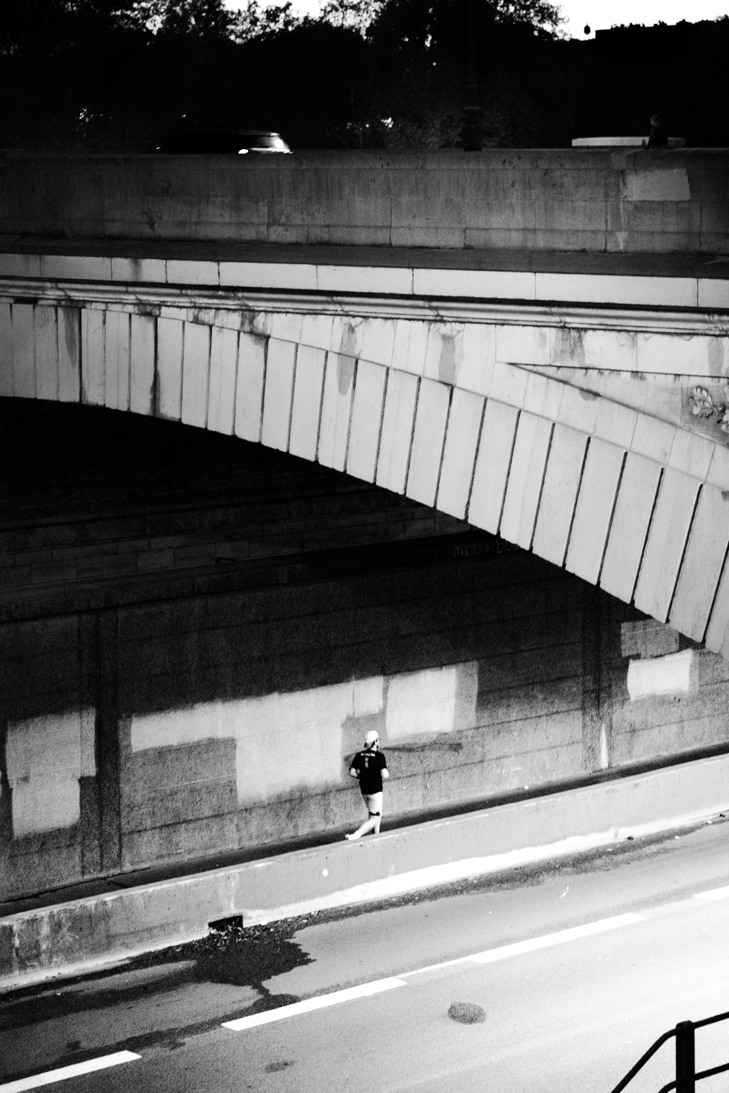 The image shows a dimly lit urban environment, with a person walking alone down a set of concrete steps in the foreground. The steps are flanked by a concrete wall with a series of horizontal beams running along the top. The background is dark, with some trees and buildings visible in the distance. The overall scene has a stark, moody atmosphere conveyed through the use of high contrast black and white tones.