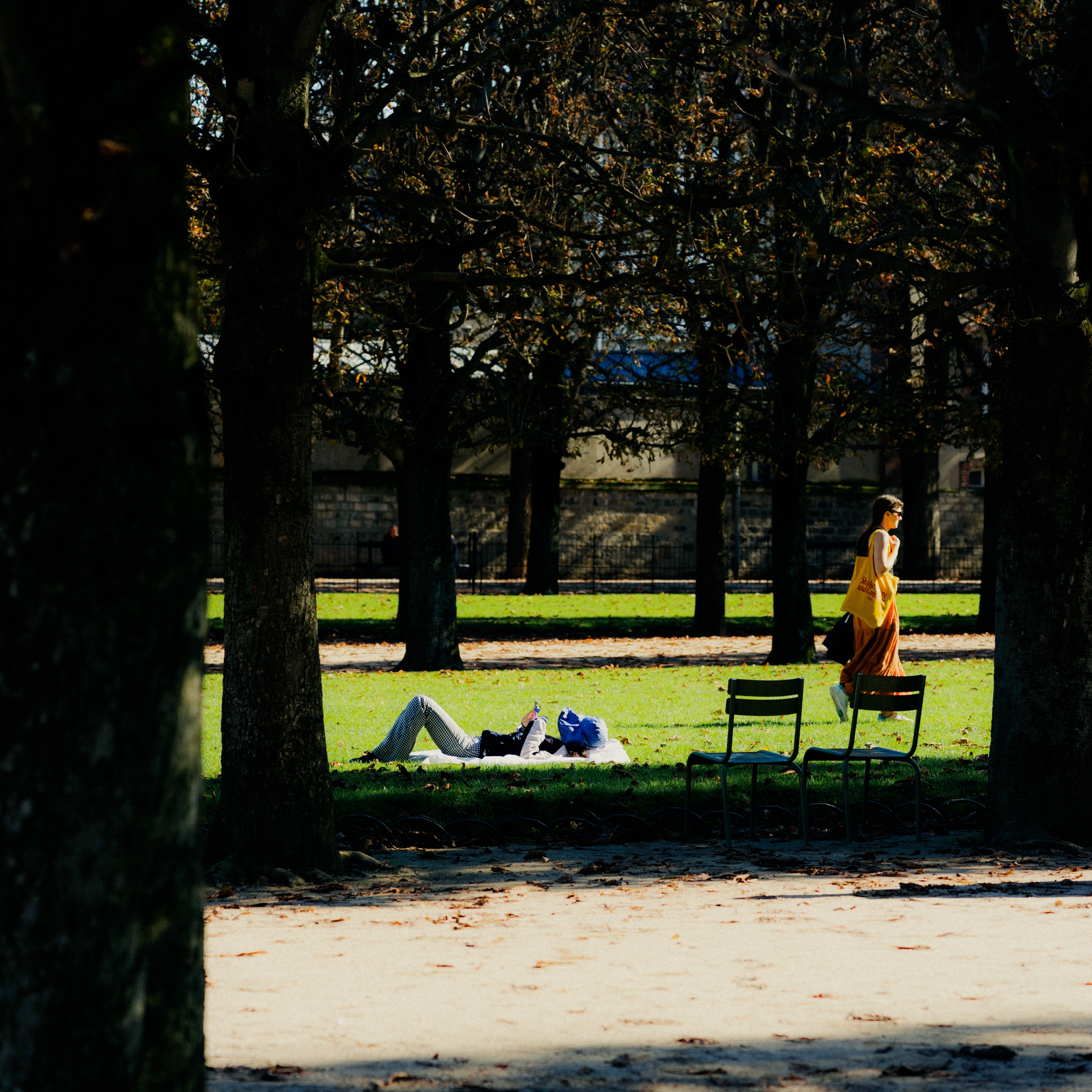 The image shows an urban park setting with a path surrounded by trees and foliage. In the foreground, a person is lying on the grass, seemingly resting or relaxing. A person in a yellow dress is standing nearby, facing the other direction. The scene has a peaceful, tranquil atmosphere.