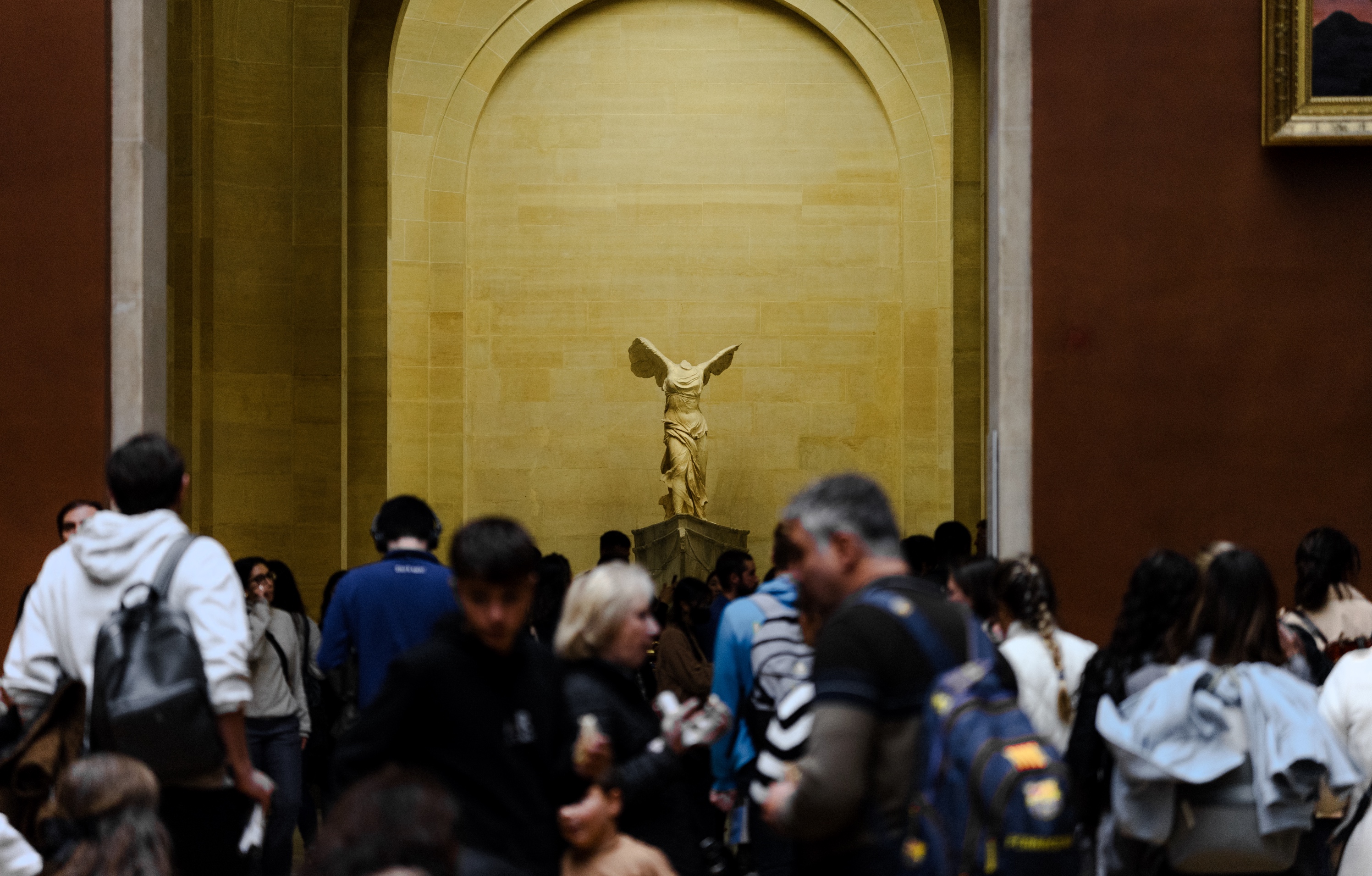 The image captures the grand and dramatic display of the Nike of Samothrace sculpture within the Louvre museum. The winged goddess of victory stands atop a pedestal, her flowing drapery and outstretched wings creating a sense of dynamic movement and power. In the foreground, a crowd of museum visitors reverently gathers to admire this famous Hellenistic masterpiece, their silhouettes contrasting against the sculpture's imposing scale and the museum's ornate stone architecture. The lighting casts a warm, golden glow on the statue, further highlighting its sculptural and artistic brilliance. The scene conveys a strong sense of the Louvre as a prestigious cultural institution preserving and showcasing some of humanity's greatest artistic achievements.