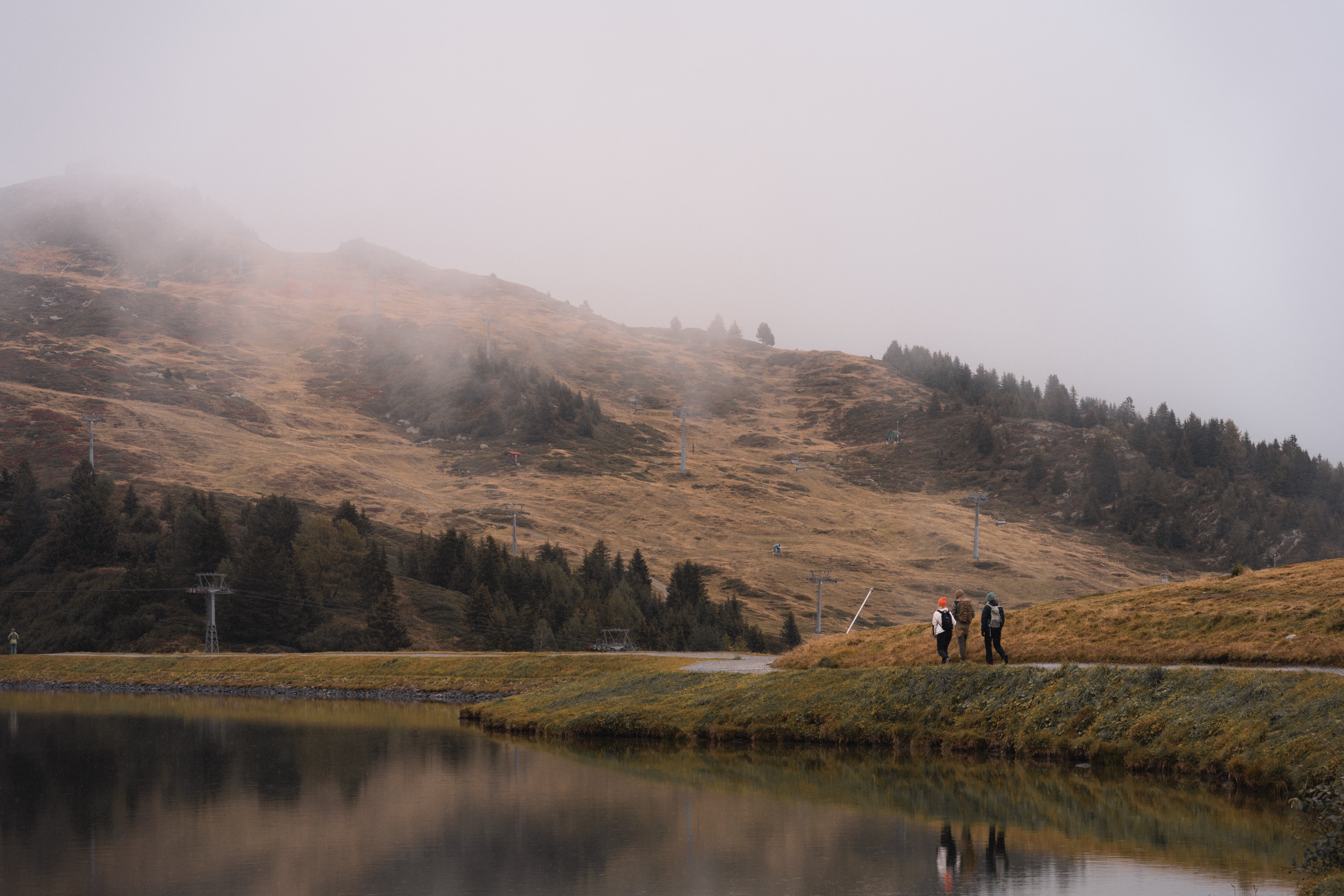 Three hikers walk along a grassy path beside a calm lake, with their reflections visible in the water. They are surrounded by an expansive mountain landscape, partially obscured by mist that hangs over the slopes. The hillside is dotted with trees and hints of autumn colors, while ski lift structures stand quietly in the background. The scene feels tranquil and remote, capturing the quiet beauty of nature in a foggy, mountainous area.