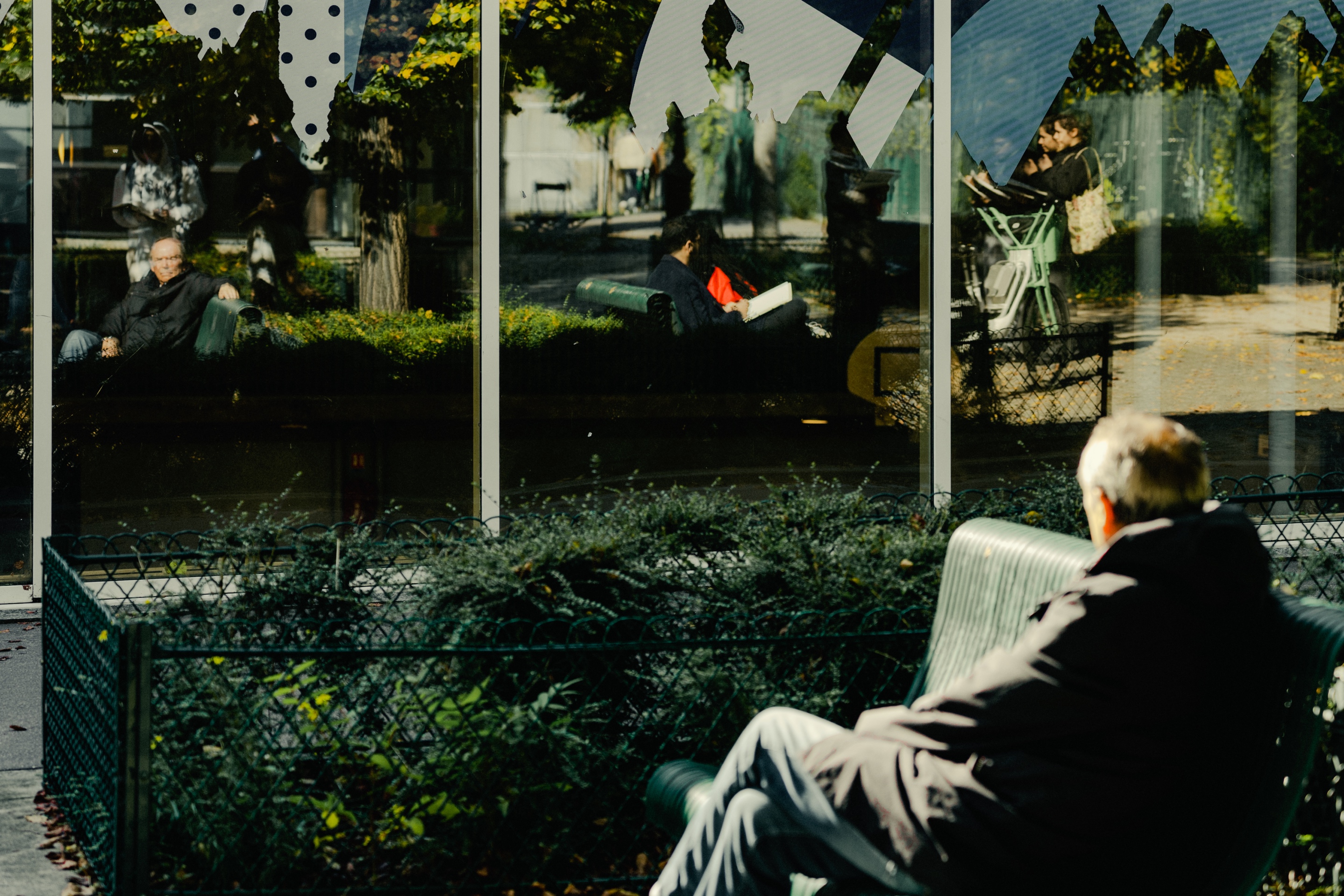 A creative shot showing both a person sitting on a park bench and their reflection in a window behind them, creating a mirror-like double image. The scene is set in what appears to be a city park or garden area with green fencing and foliage. Polka dot decorations are visible at the top of the window panels, and the lighting suggests it's during daytime. The composition creates an interesting interplay between the real person and their reflection.