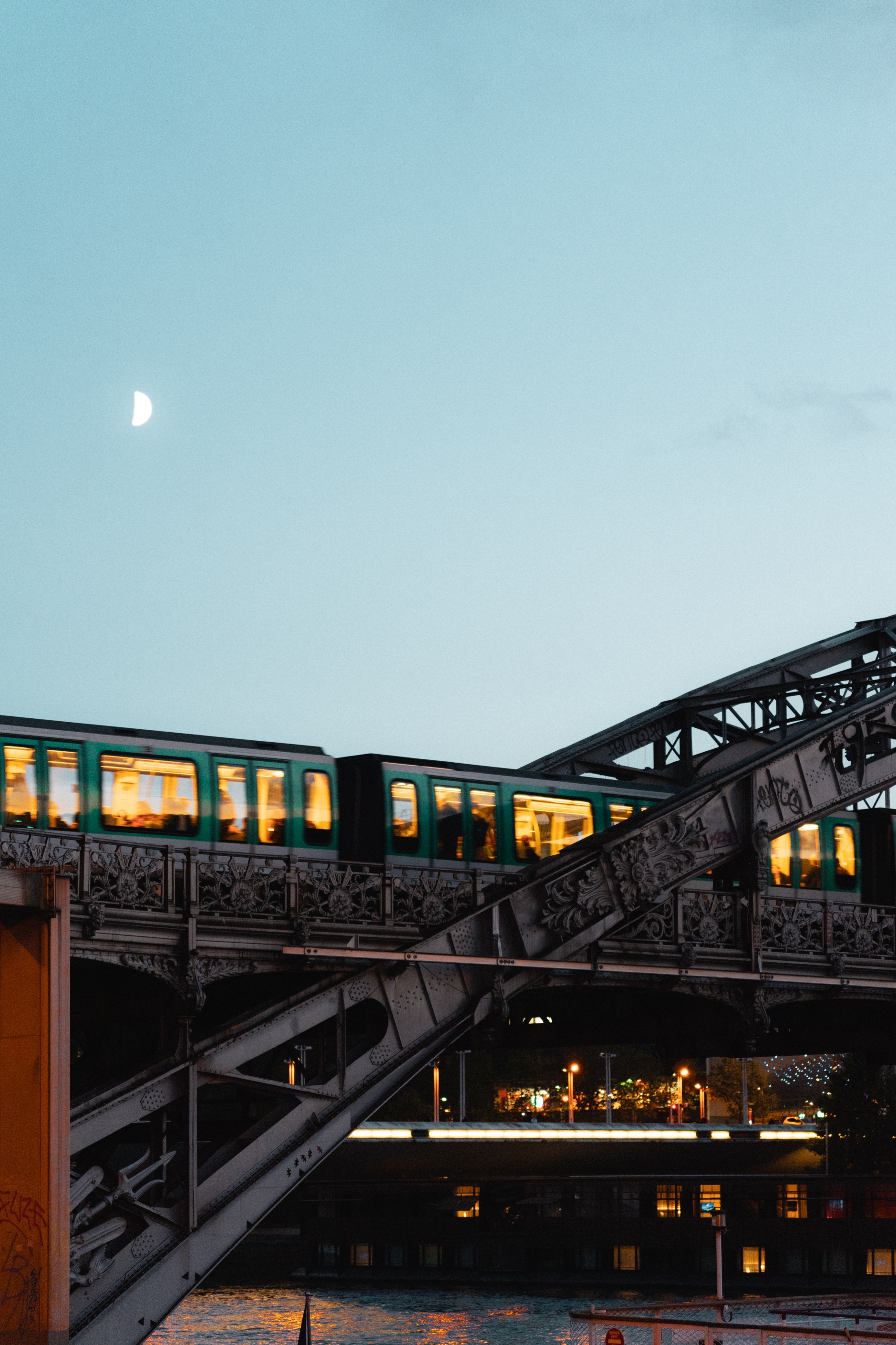 A twilight scene of a Paris Metro train crossing an ornate iron bridge, likely part of the elevated Metro system. The green train cars are lit from within, creating a warm golden glow through their windows. A half moon is visible in the pale blue sky above. Below the bridge, city lights reflect on what appears to be the Seine River. The bridge's detailed iron latticework showcases classic Parisian architectural design.