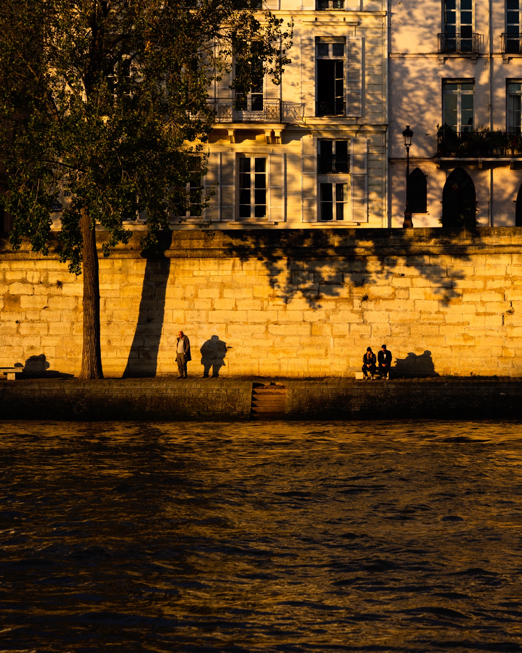 The image depicts a serene scene along the banks of the Seine River in Paris. The historic buildings lining the riverfront are bathed in the warm, golden light of the late afternoon sun, casting long shadows across the river. A few people can be seen sitting on the stone steps leading down to the water, enjoying the peaceful atmosphere. The overall scene evokes a sense of timeless elegance and tranquility found along the iconic Seine River in the French capital.